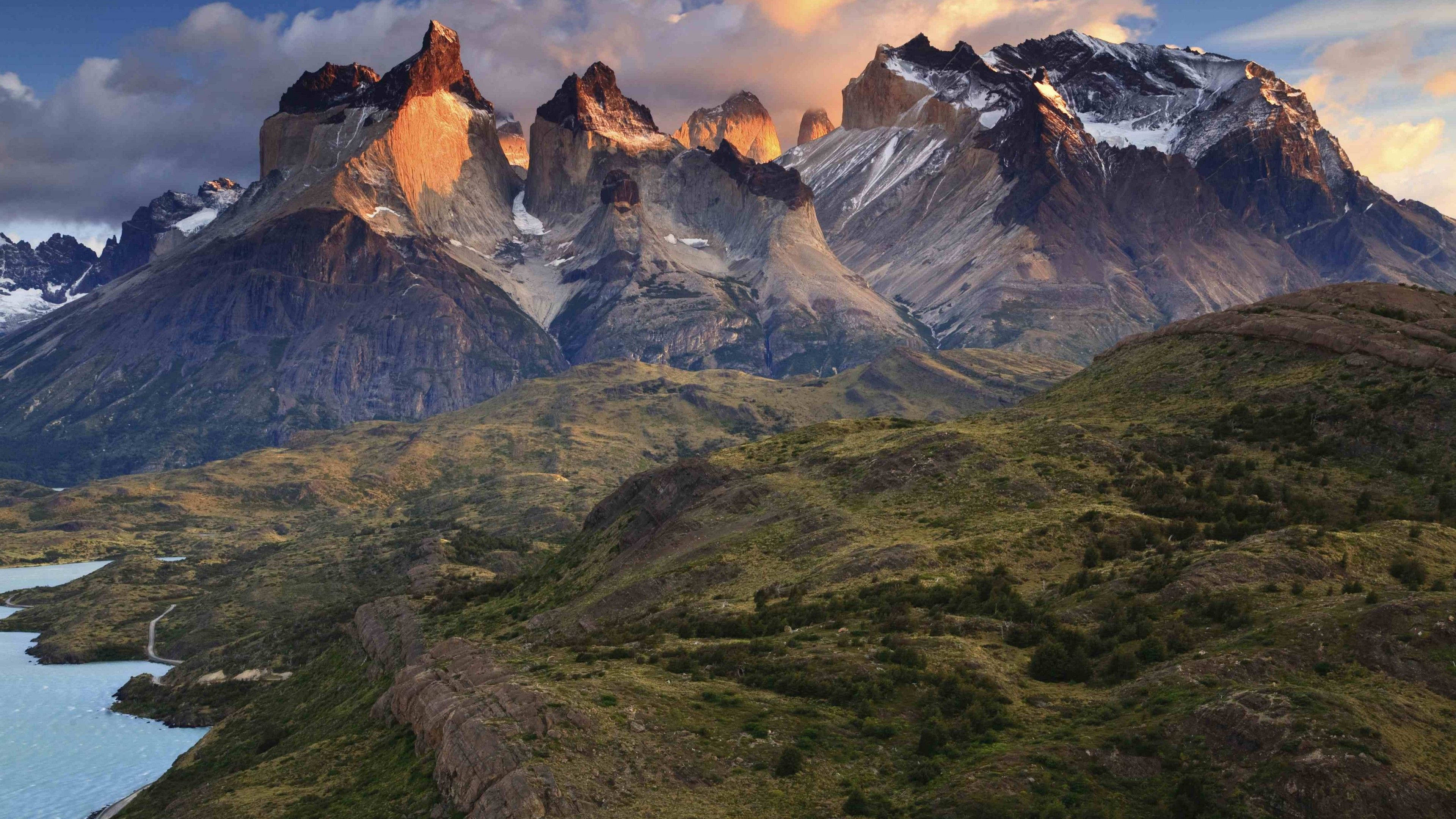 Patagonia 桌布 壁紙 — 夕陽將鋸齒狀的雪山山峰沐浴在金色光芒中，下方是寧靜的湖泊和起伏的綠色山丘。