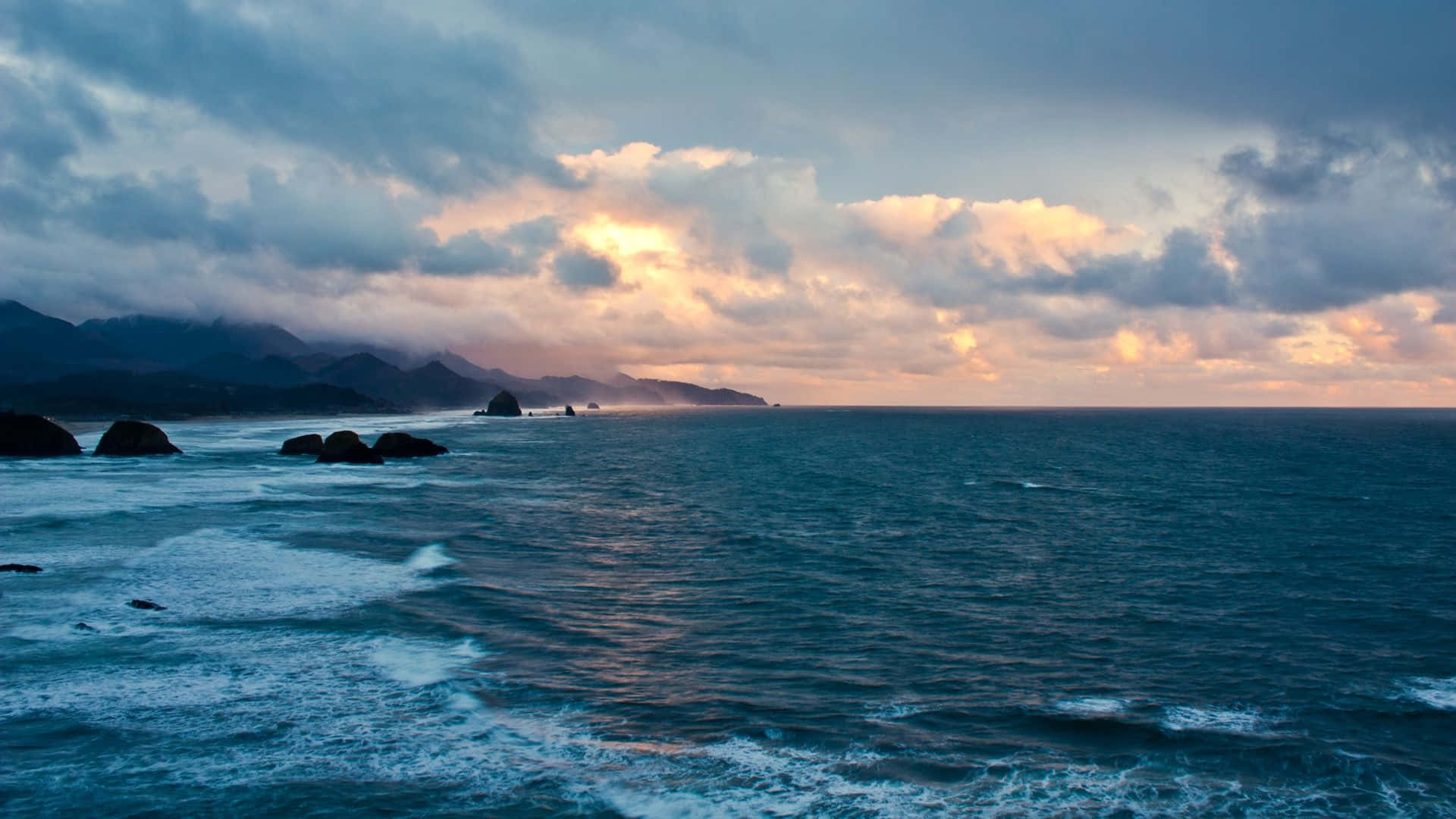Beautiful Ocean 桌布 壁紙 — 夕陽將雲彩染成金色，映照在波濤洶湧的海洋上，暗黑的岩石與遠山在陰鬱的天空下若隱若現。