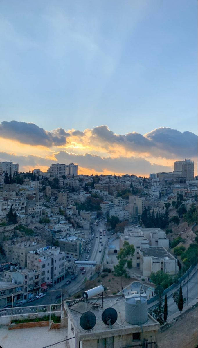 Jordan 桌布 壁紙 — 夕陽將雲彩染成金色，籠罩著一座密集的山丘城市，蜿蜒的道路和屋頂水箱清晰可見。