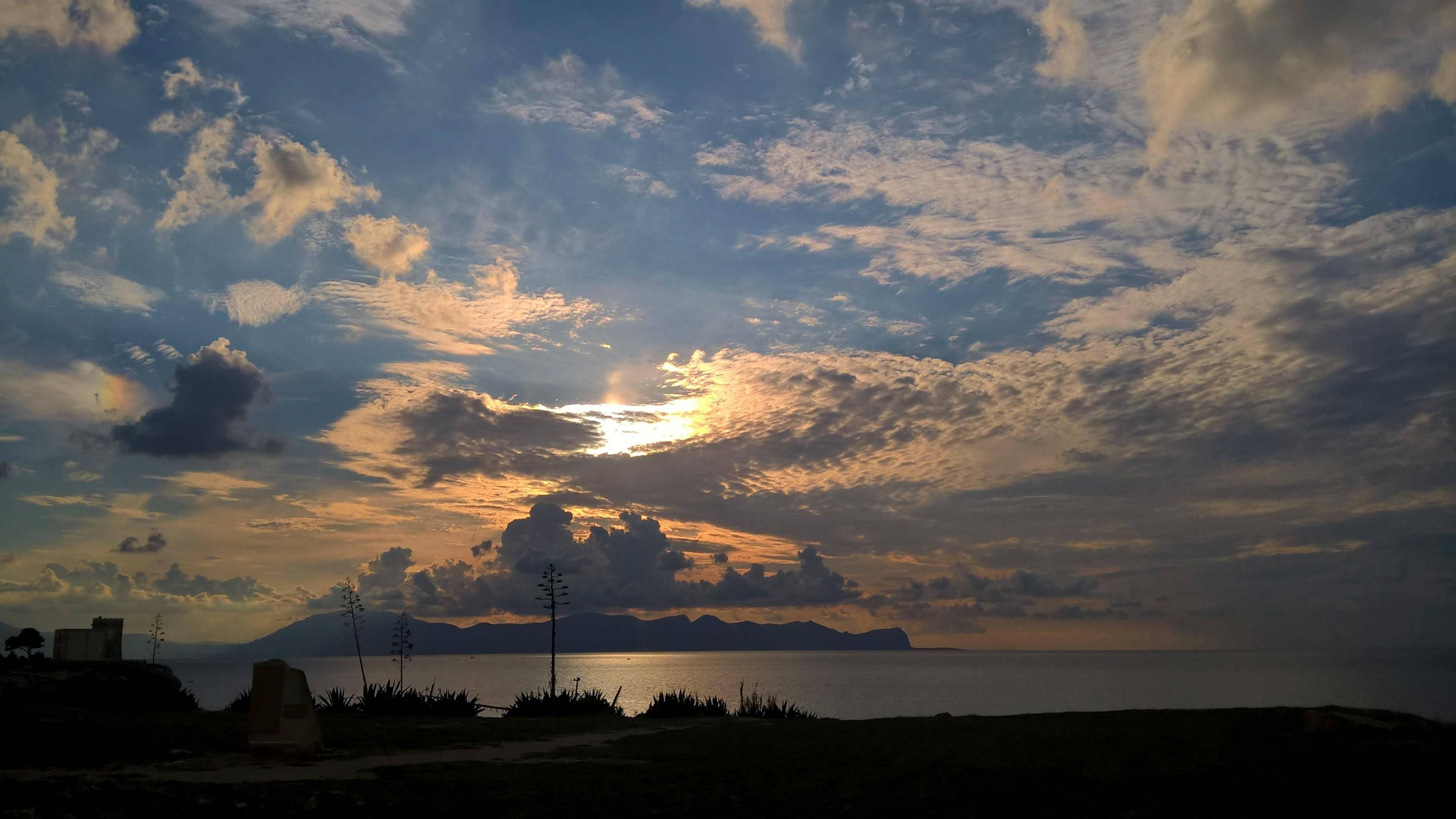 天堂 桌布 壁紙 — 夕陽將雲彩染成金色和藍色，映照在平靜的海面上，遠處有山巒的輪廓。