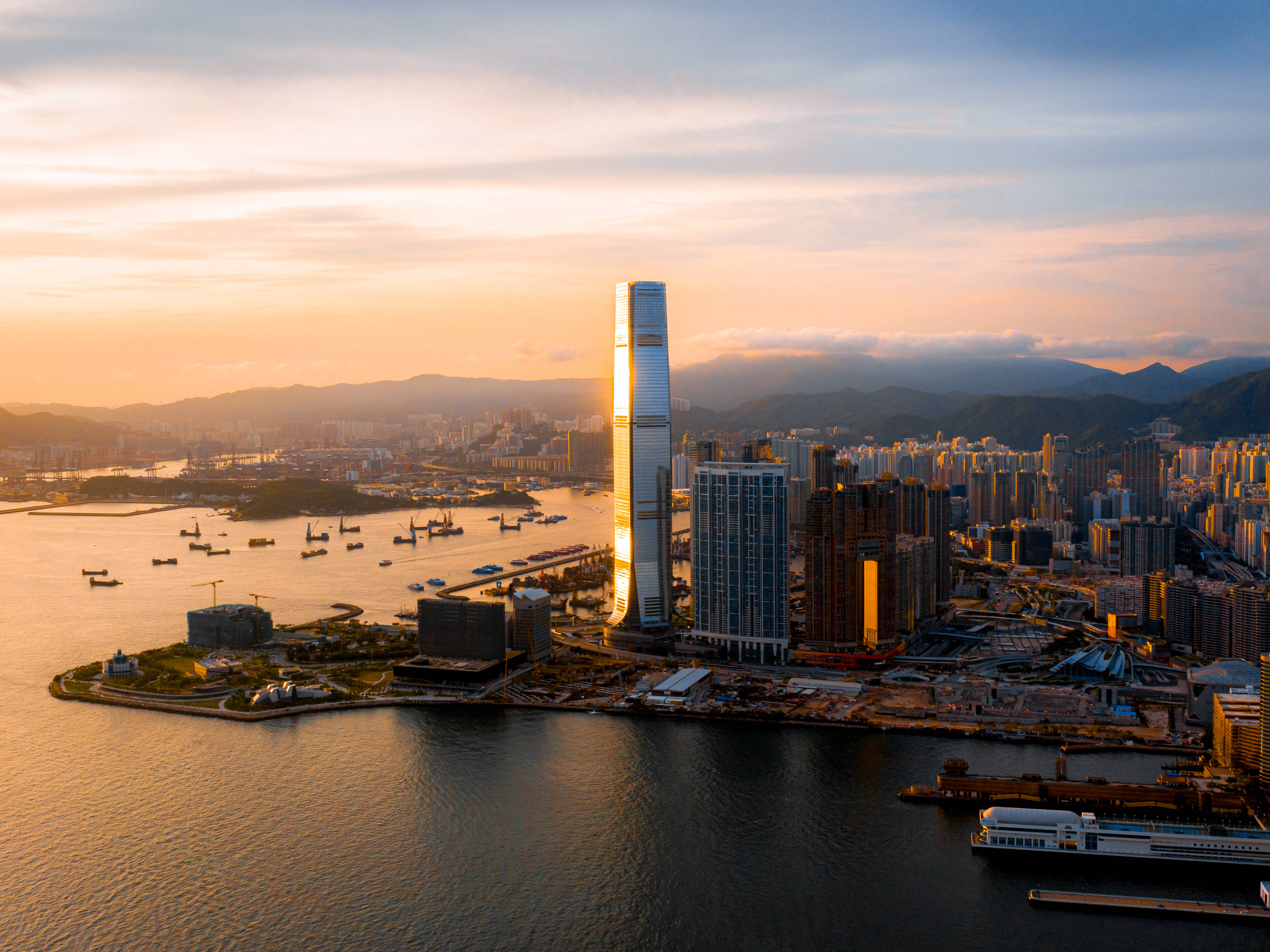 香港 桌布 壁紙 — 夕陽將香港天際線沐浴在金色光芒中，摩天大樓倒映在點綴著船隻的平靜水面上。