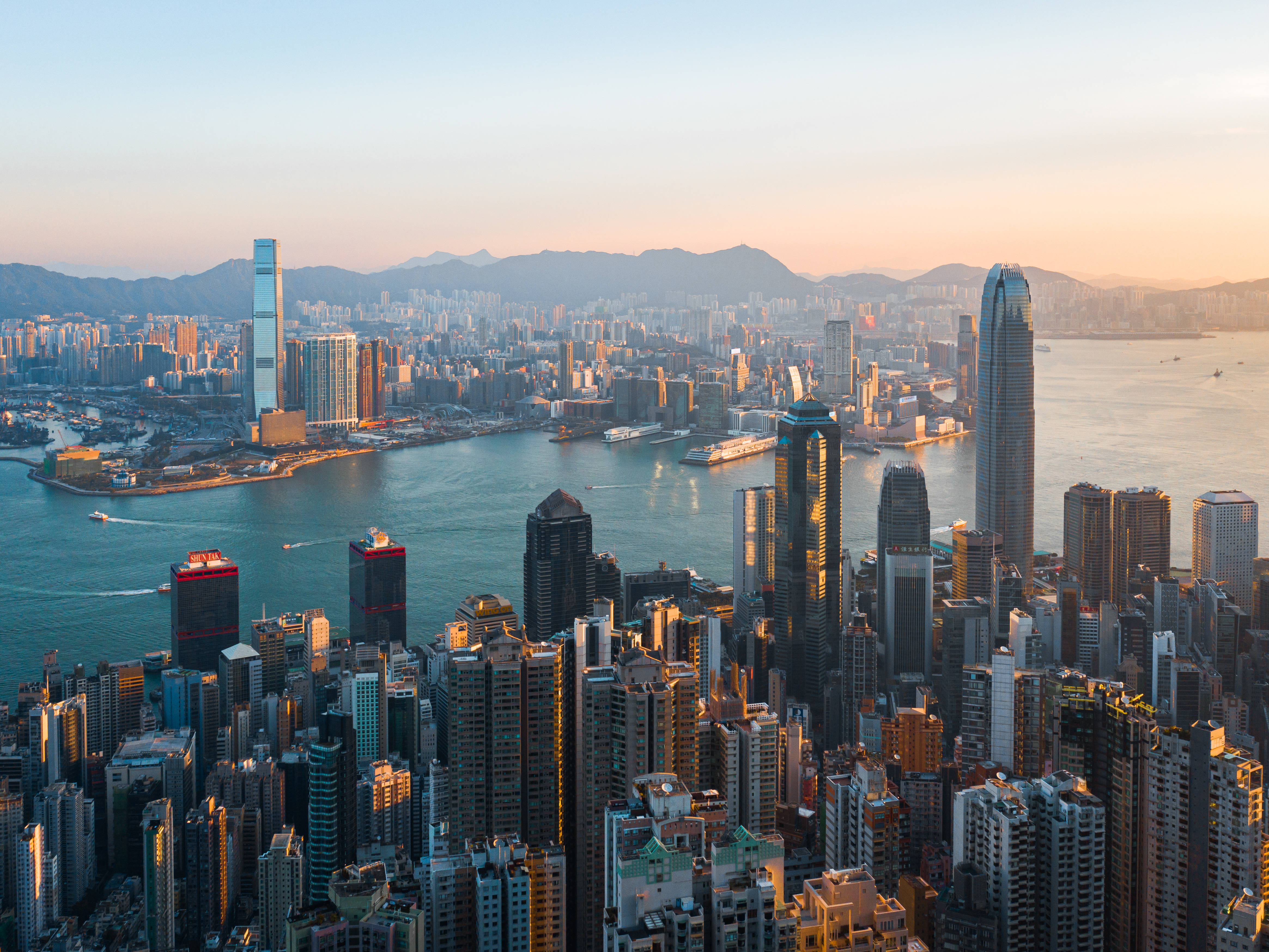 香港 桌布 壁紙 — 夕陽將香港天際線沐浴在金色光芒中，摩天大樓聳立在維多利亞港之上，遠山在柔和的薄霧中。