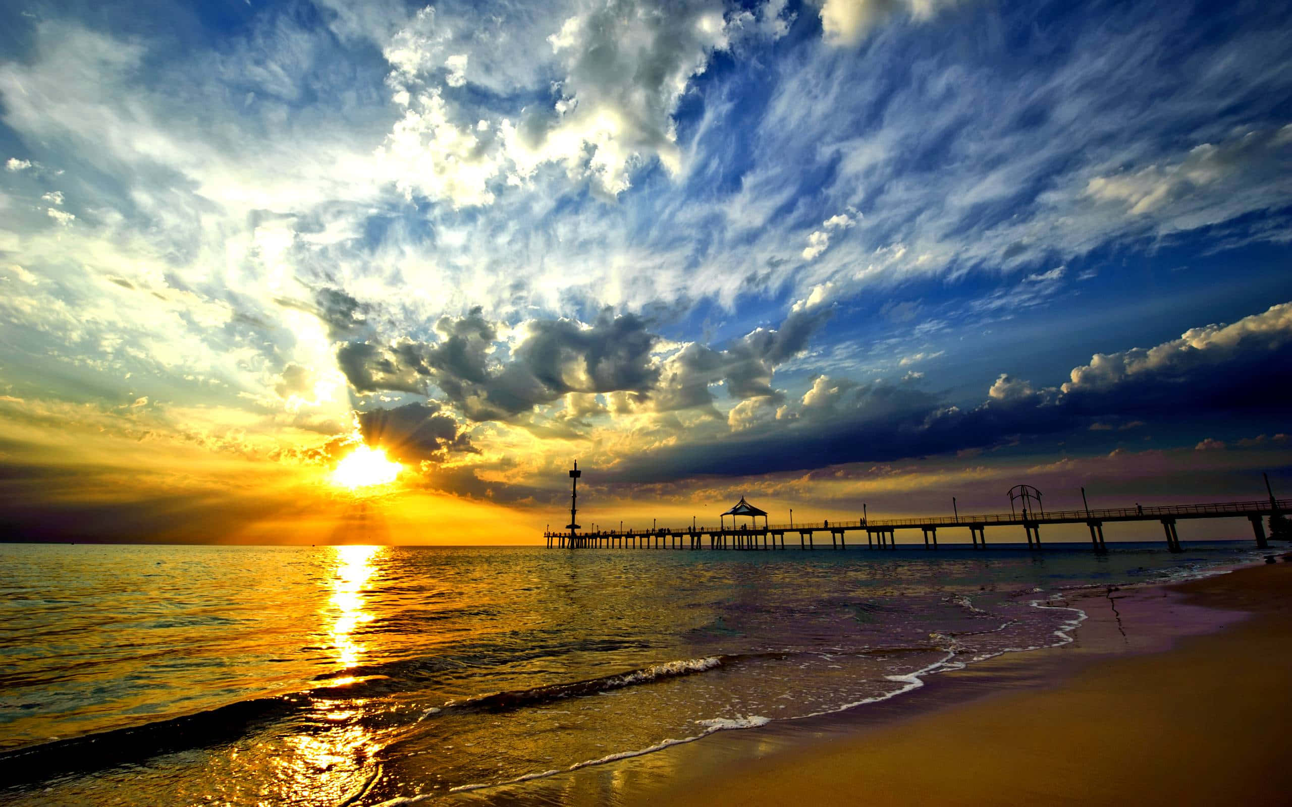 Beautiful Sky 桌布 壁紙 — 夕陽映照海洋，金色光芒在波浪上閃爍，長長的碼頭在戲劇性的雲層下延伸至地平線。