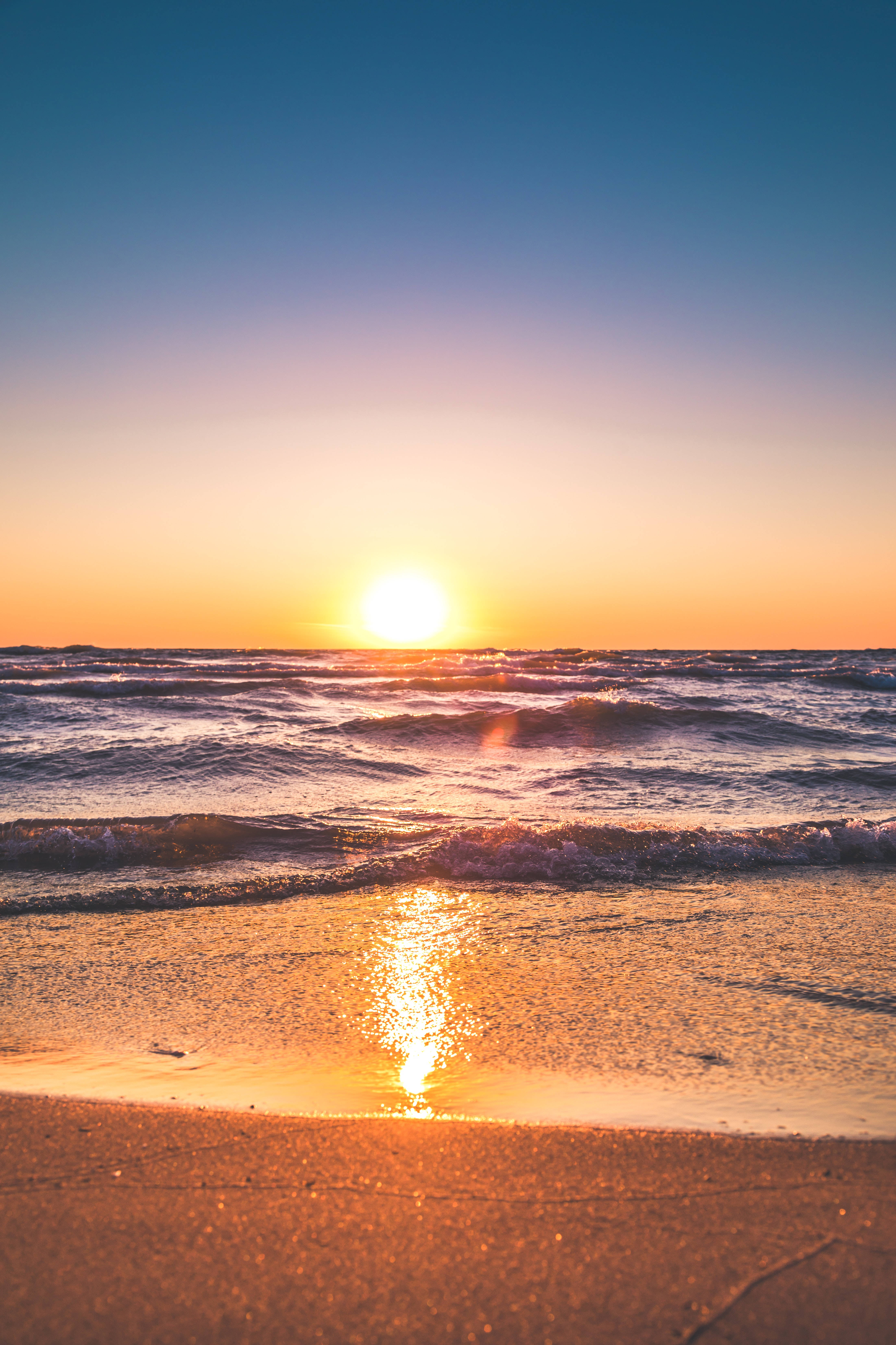 夏天 iPhone 桌布 壁紙 — 夕陽映照海洋，金色波浪拍打沙灘，天空呈現漸變色彩。