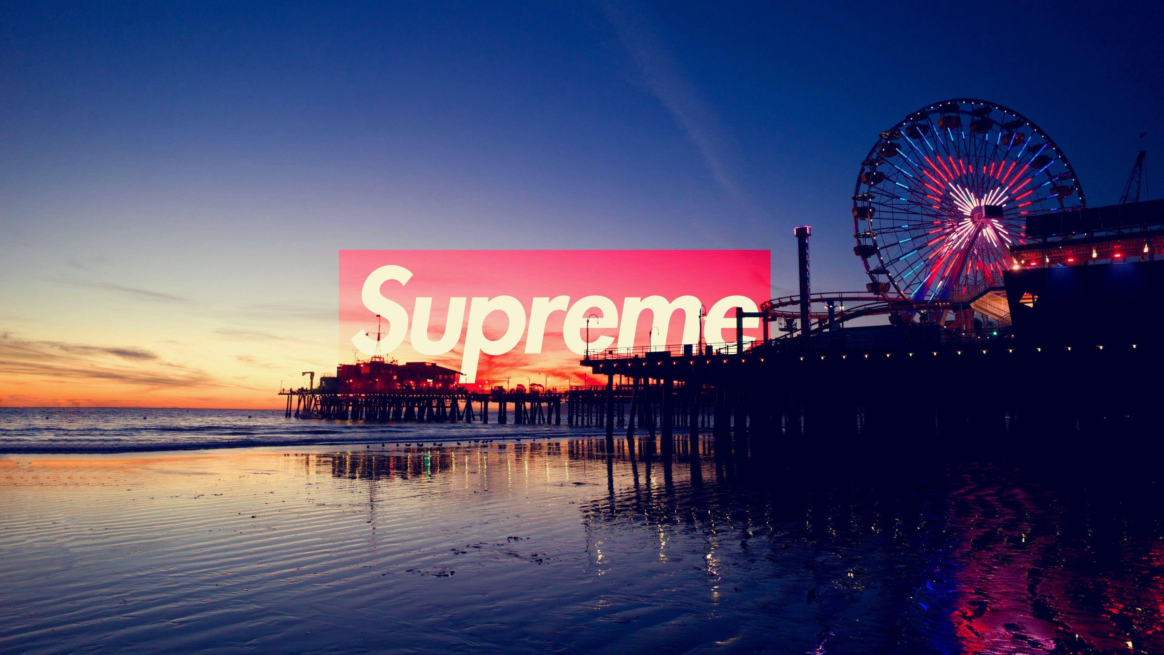 supreme 桌布 壁紙 — 夕陽映照海灘，燈光點亮的碼頭和摩天輪倒映在濕沙上，並覆蓋著粉紅色的Supreme標誌。