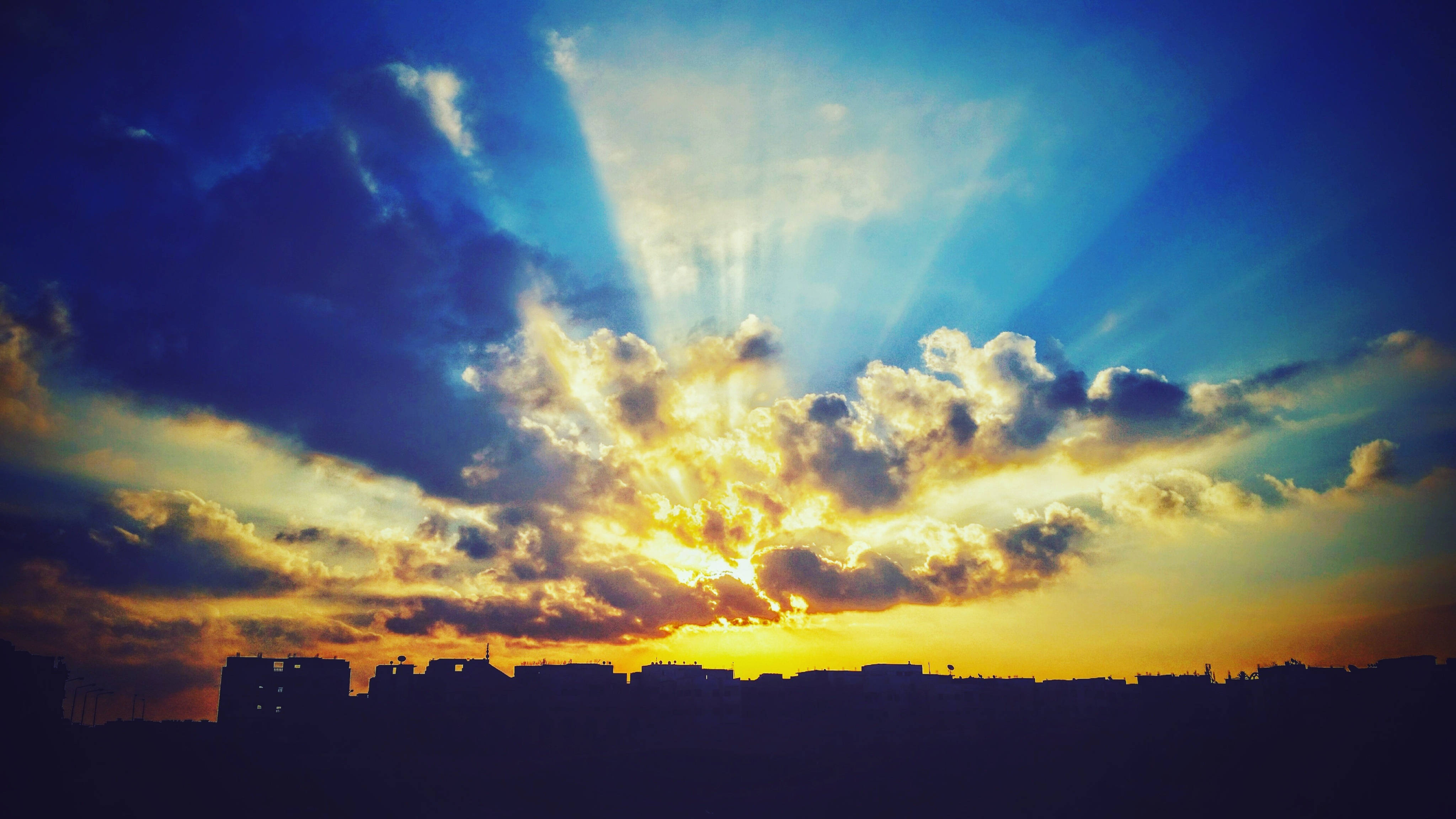 天空 桌布 壁紙 — 夕陽時分，陽光穿透戲劇性的雲層，照射在城市天際線之上，在深藍色的天空中投下金色的光芒。