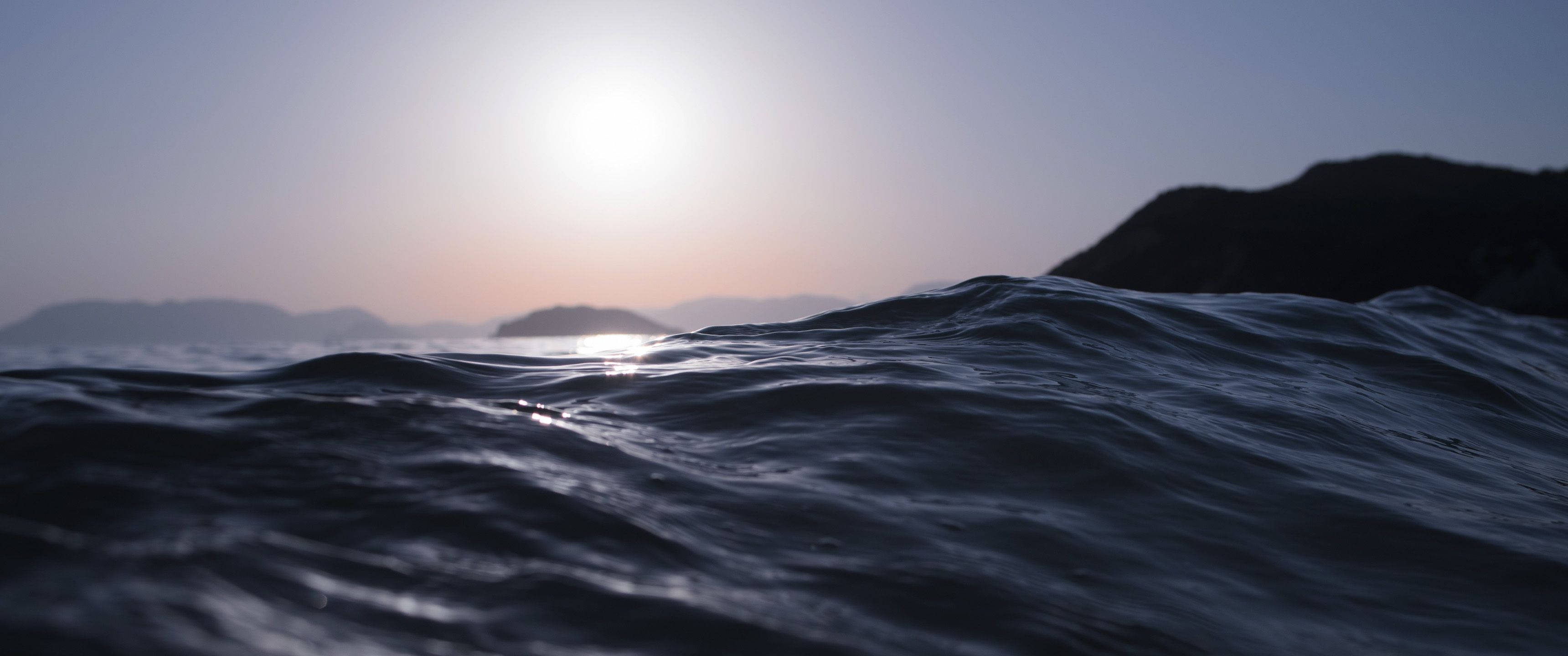 3840x1600 桌布 壁紙 — 夕陽柔和地照耀在深色海浪上，遠方山丘在漸暗的天空下形成剪影。