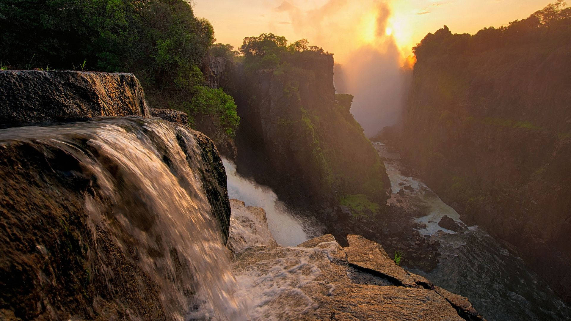 Zimbabwe 桌布 壁紙 — 夕陽沐浴著咆哮的瀑布，瀑布穿過深峽谷，兩旁是鬱鬱蔥蔥的綠色懸崖。