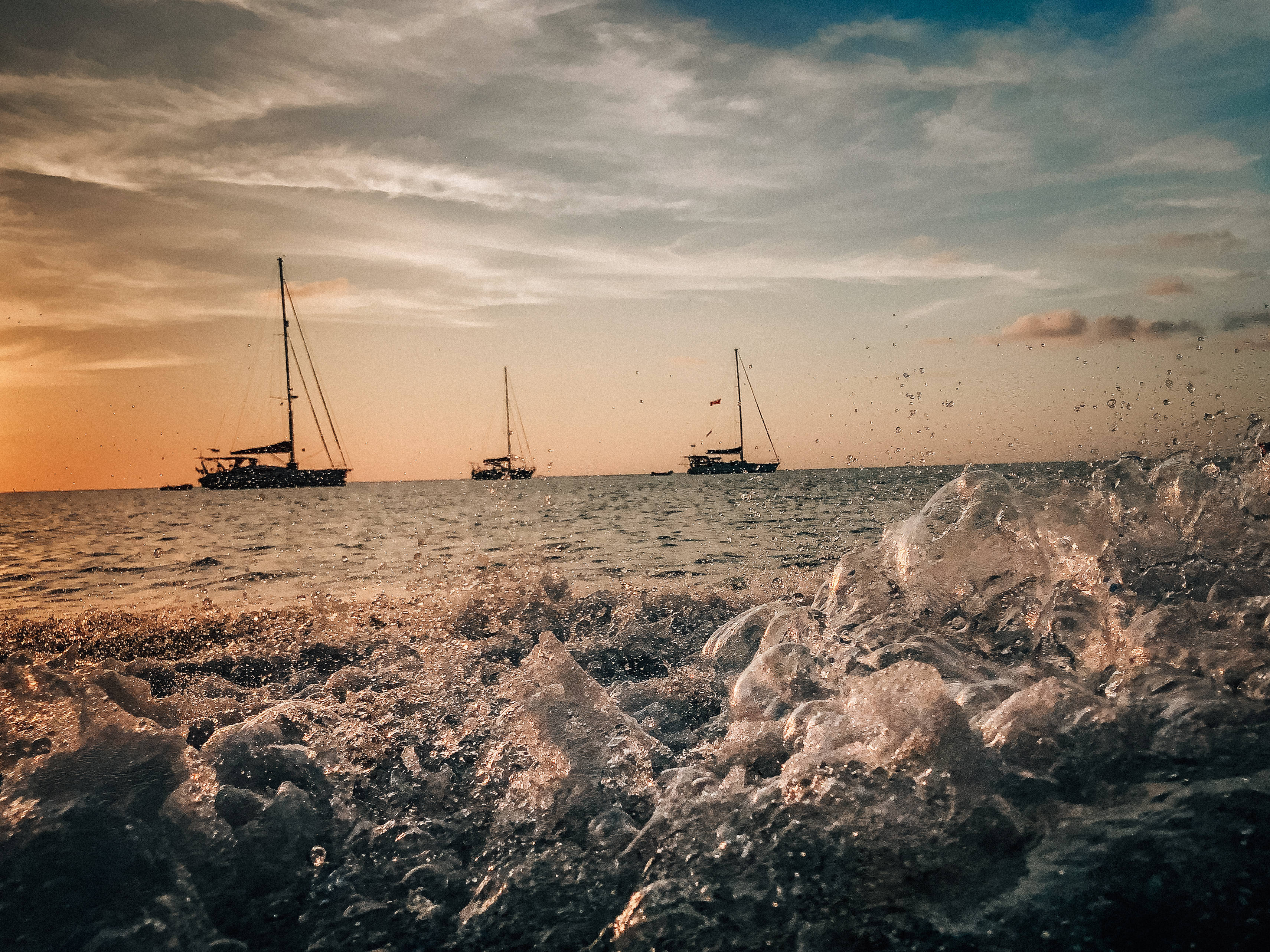 Honduras 桌布 壁紙 — 夕陽海浪在前景拍打，三艘帆船在金色天空下平靜的海面上滑行。