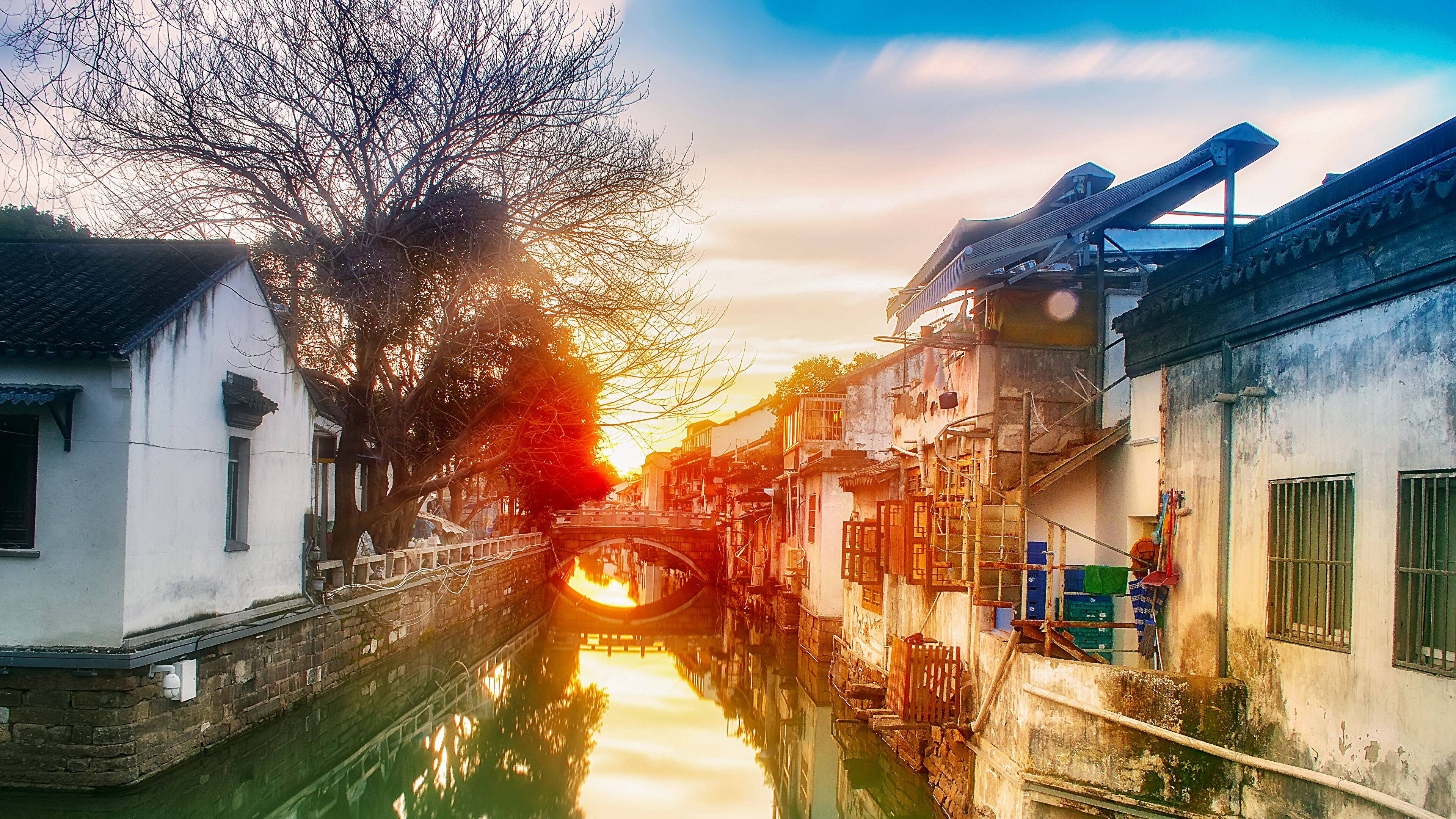 Suzhou 桌布 壁紙 — 夕陽溫暖地照耀在一條寧靜的運河上，兩旁是傳統的白色牆壁建築和光禿禿的樹木。