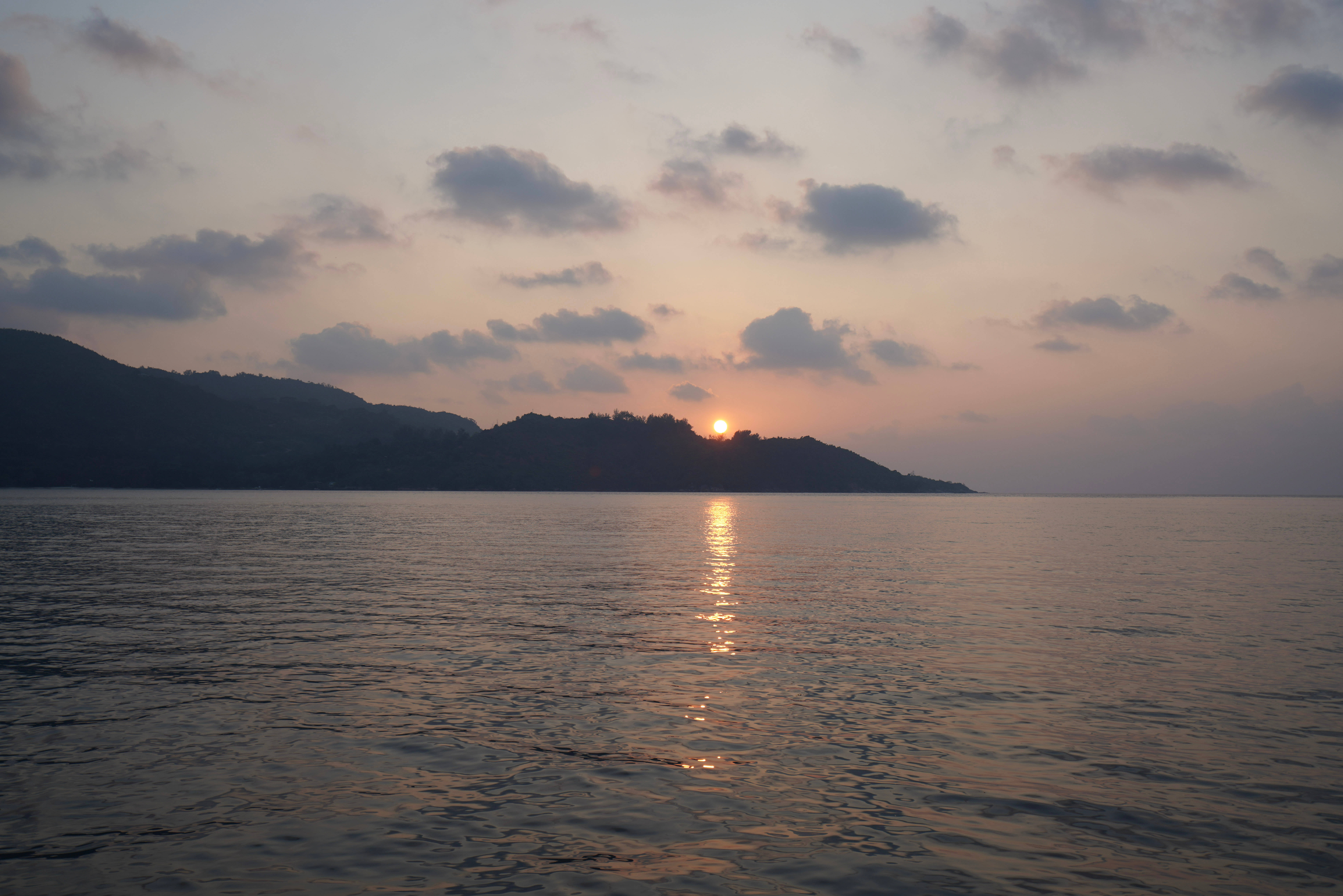 Seychelles 桌布 壁紙 — 夕陽溫暖地照耀在平靜的水面上，遠山在柔和的粉紅色和橙色雲彩中形成剪影。