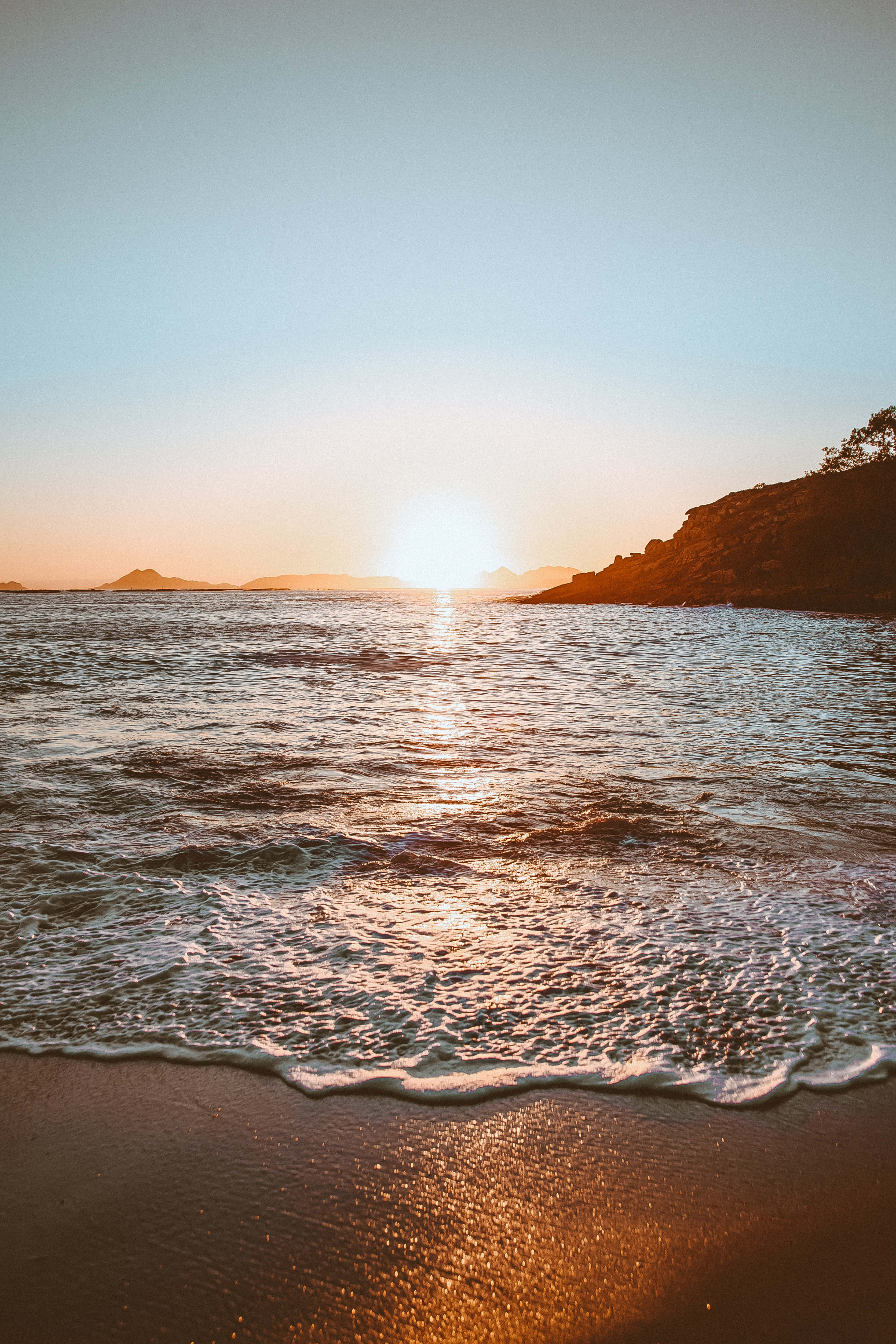 日出 桌布 壁紙 — 夕陽溫暖地照耀著海浪拍打沙灘，遠方的懸崖和島嶼在金色光線中形成剪影。