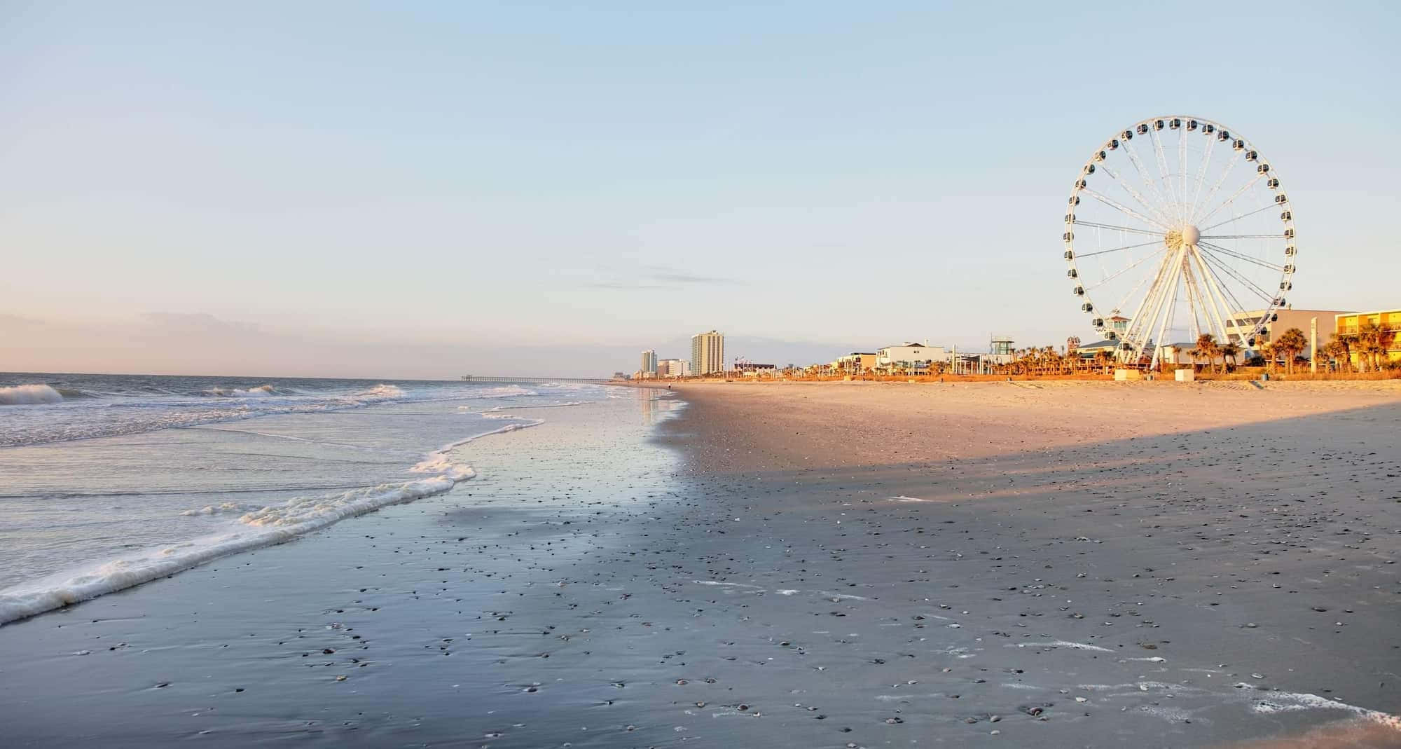 Myrtle Beach 桌布 壁紙 — 夕陽灑在沙灘上，伴隨著輕柔的海浪，一個巨大的白色摩天輪高高矗立在岸邊。