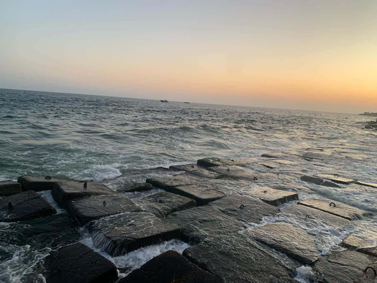 Sun rise at Alexandria, Egypt 壁紙 — 夕陽灑落，海浪拍打深色石堤，遠方船隻在平靜海平線上。