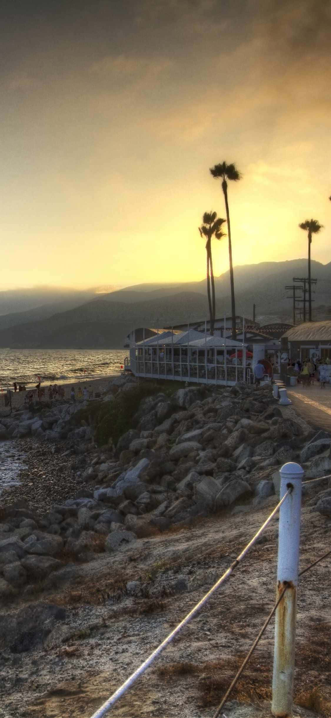 iPhone Xs Malibu 桌布 壁紙 — 夕陽灑落在岩石海岸小徑上，棕櫚樹和玻璃建築映襯著遠山。