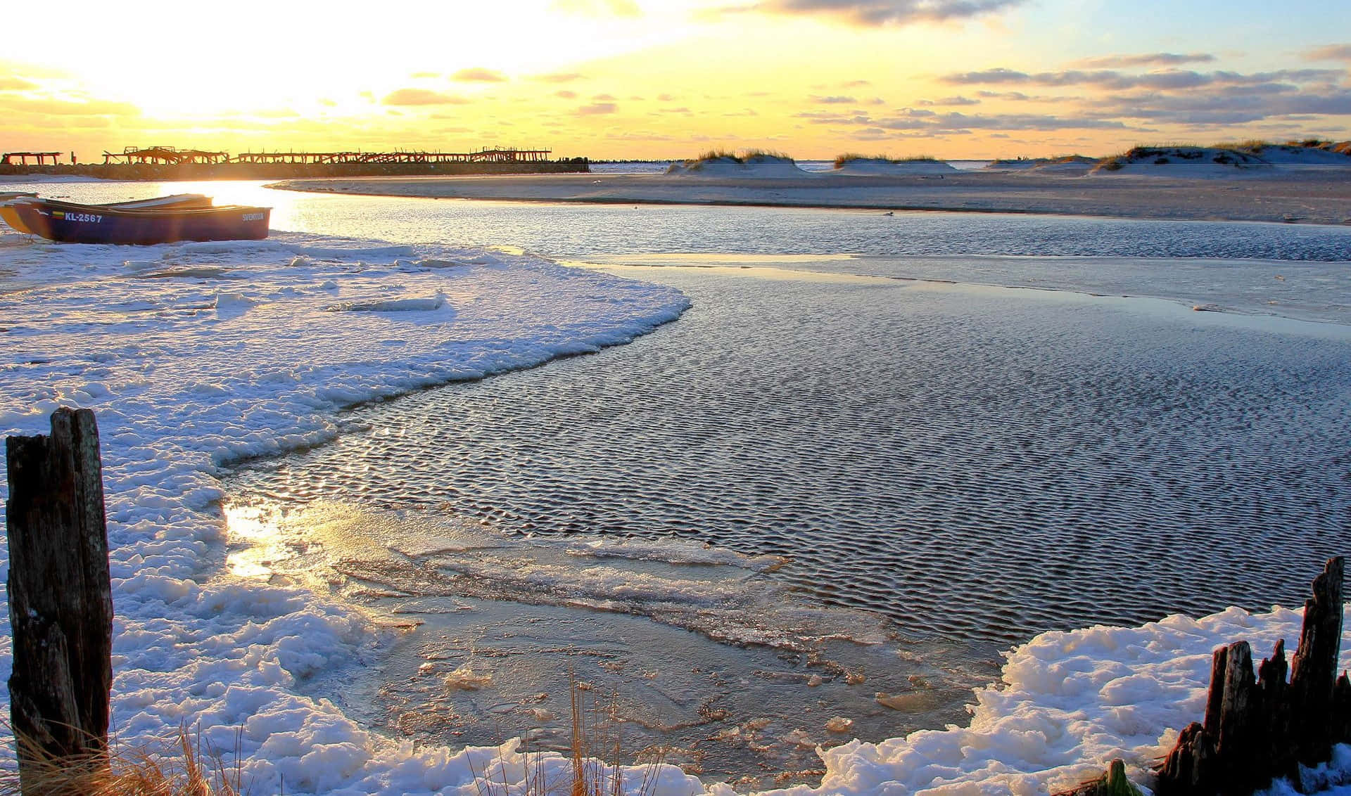 Frozen Lake 桌布 壁紙 — 夕陽照耀在冰封的水面和雪覆蓋的岸邊，一艘孤獨的小船停靠在風化的木樁旁。
