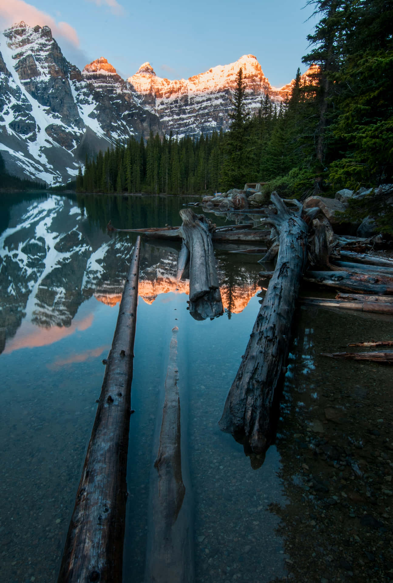 白雪覆蓋的山脈在夕陽下倒映在平靜的湖面上，前景有倒下的原木。 — 4K Android 壁紙