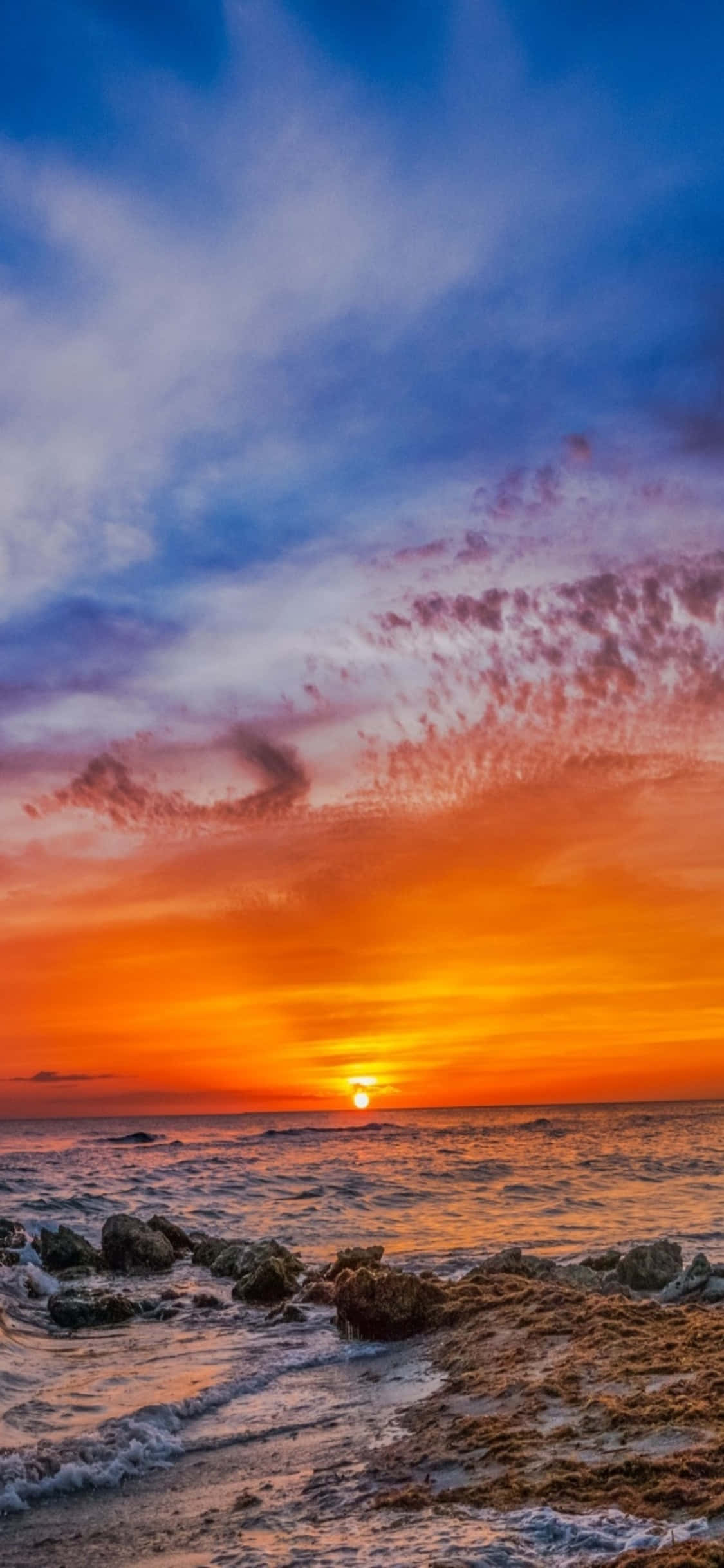 iPhone X Beach 桌布 壁紙 — 夕陽燦爛，天空染上火橙色和深藍色，海浪拍打岩石海岸。