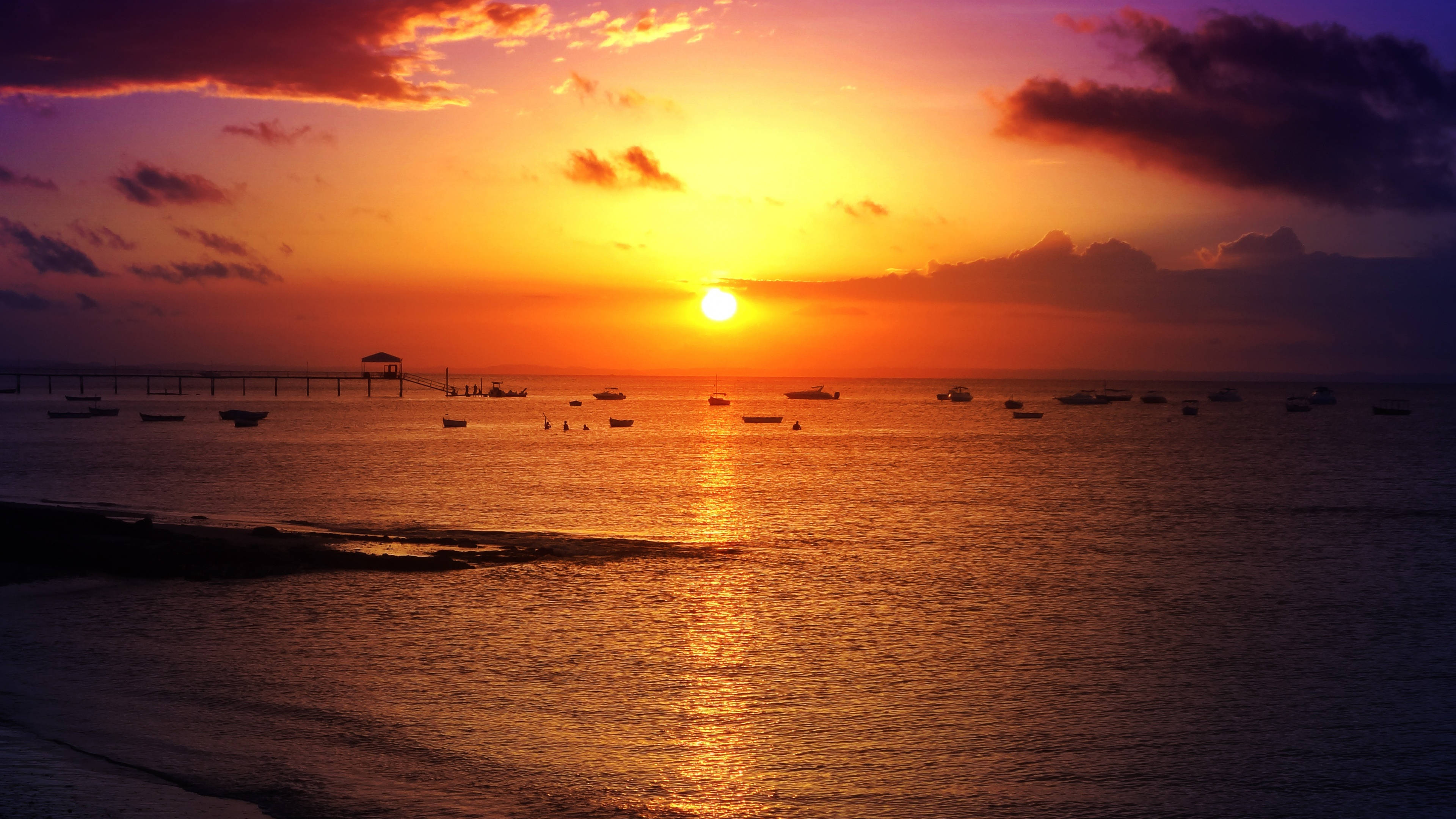 天空 桌布 壁紙 — 夕陽燦爛，天空染上熾熱的橙色和紫色，船隻在碼頭附近的平靜水面上休憩。