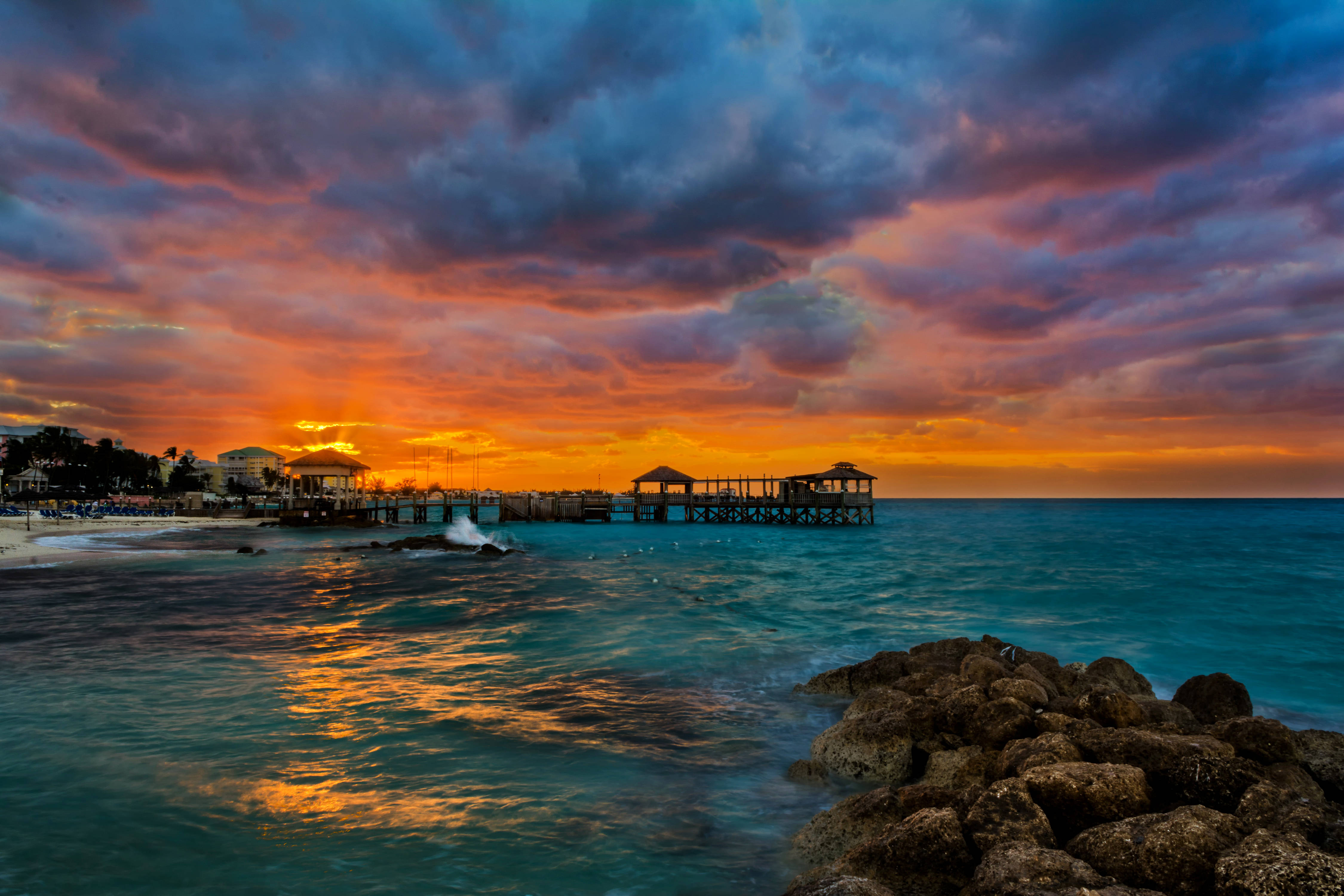 Nassau Bahamas 桌布 壁紙 — 夕陽燦爛，將天空染成火橙色和深紫色，映照在碧綠的海洋、木碼頭和岩石海岸上。
