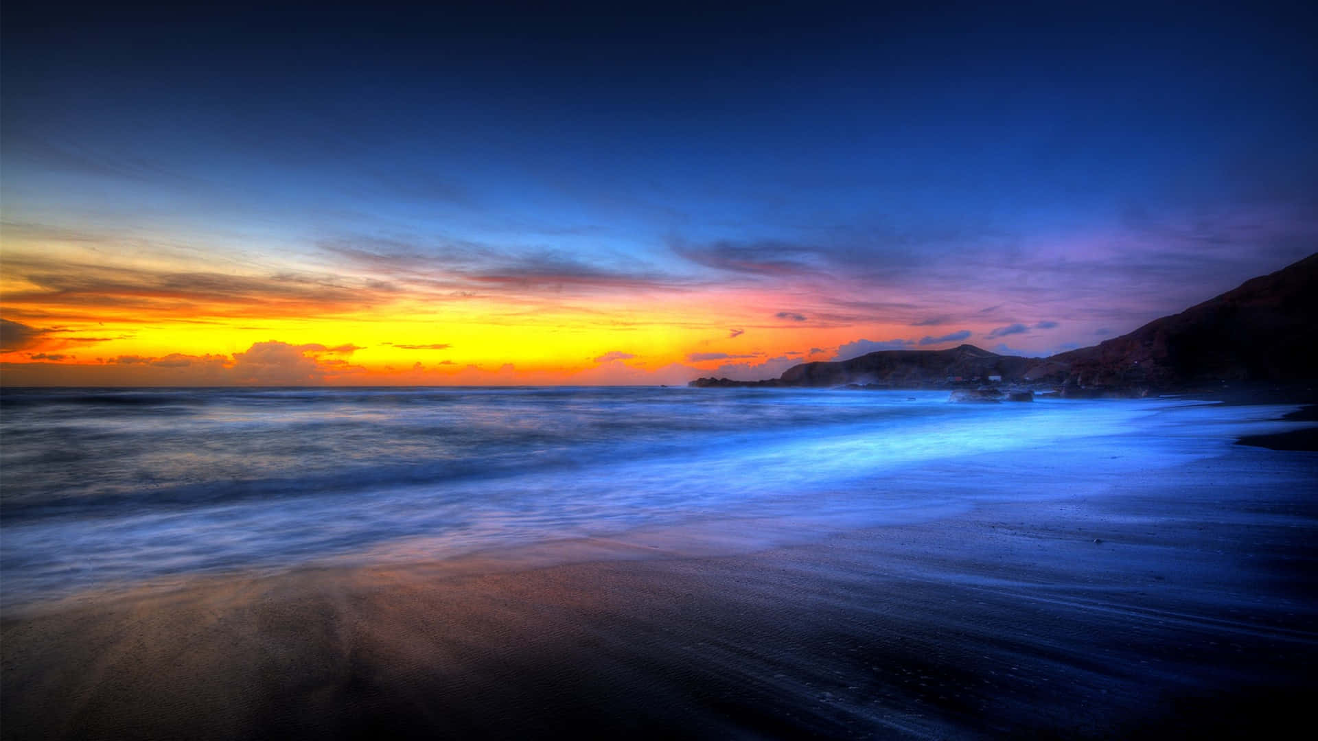 Beautiful Beach 桌布 壁紙 — 夕陽燦爛，將天空染成火橙色和深藍色，海浪輕輕拍打著深色沙灘。