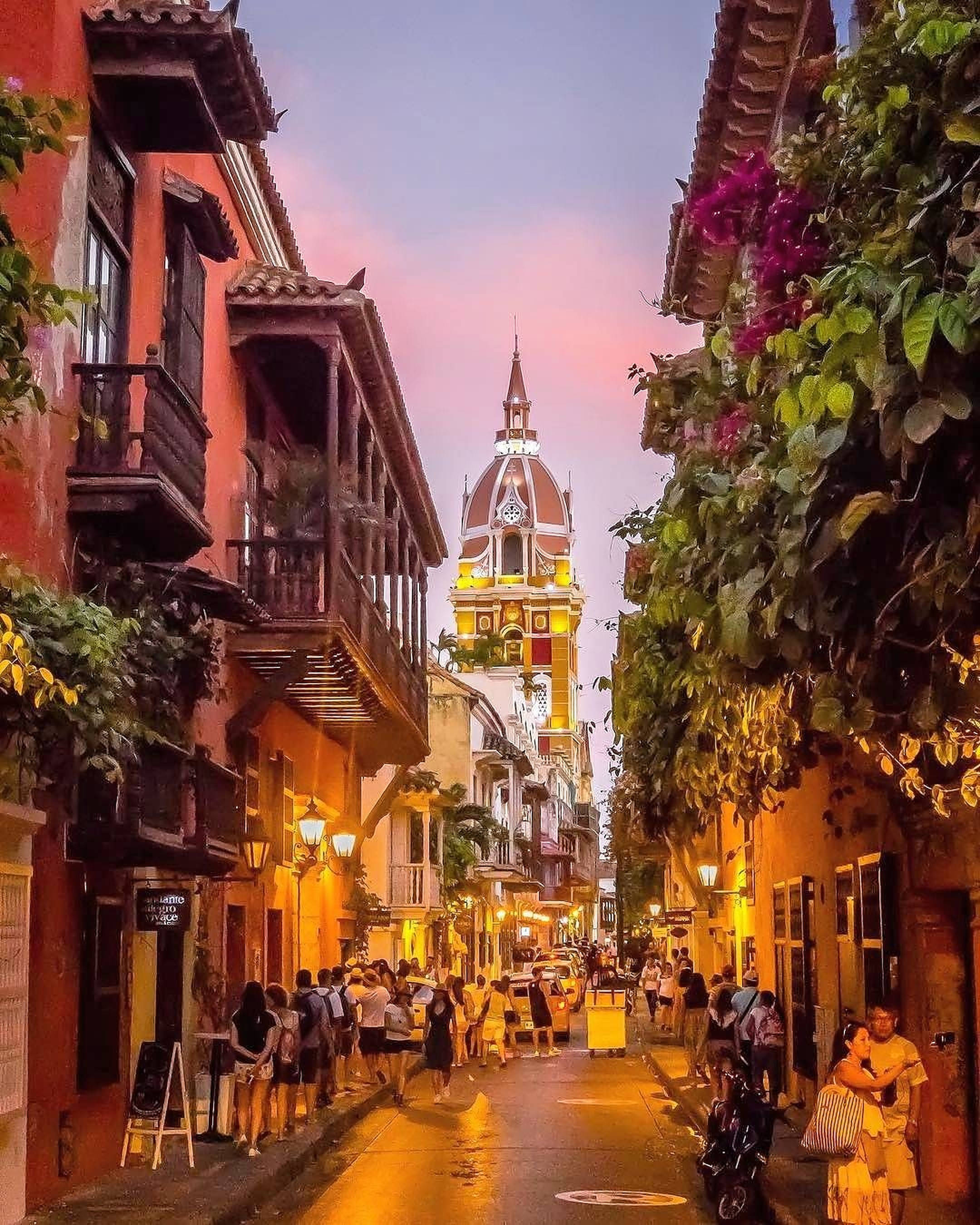Colombia 桌布 壁紙 — 夕陽燦爛，溫暖的陽光灑在一條狹窄的殖民風格街道上，人們漫步經過色彩繽紛的建築，建築上掛滿了鮮花，遠處的鐘樓閃閃發光。