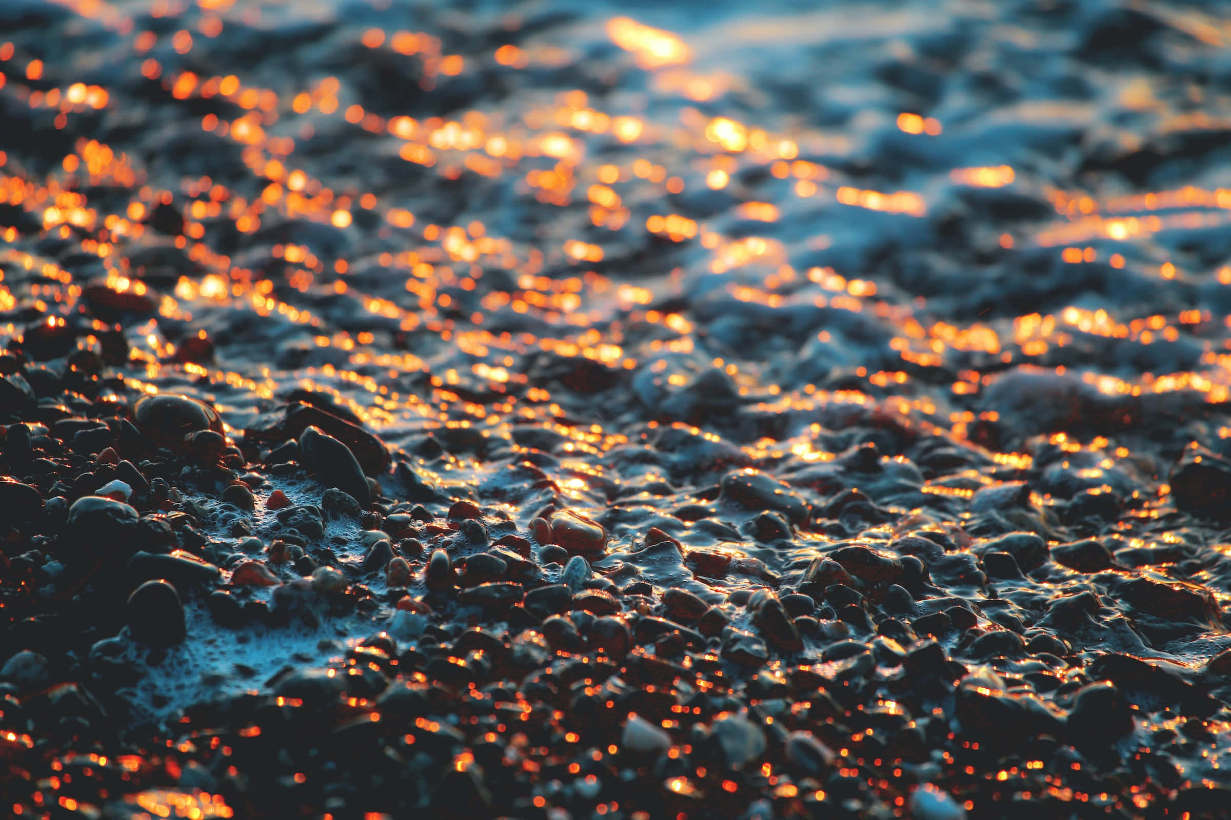海洋 桌布 壁紙 — 夕陽的光線在海灘濕潤的鵝卵石上閃爍，形成閃閃發光的金色和藍色紋理表面。