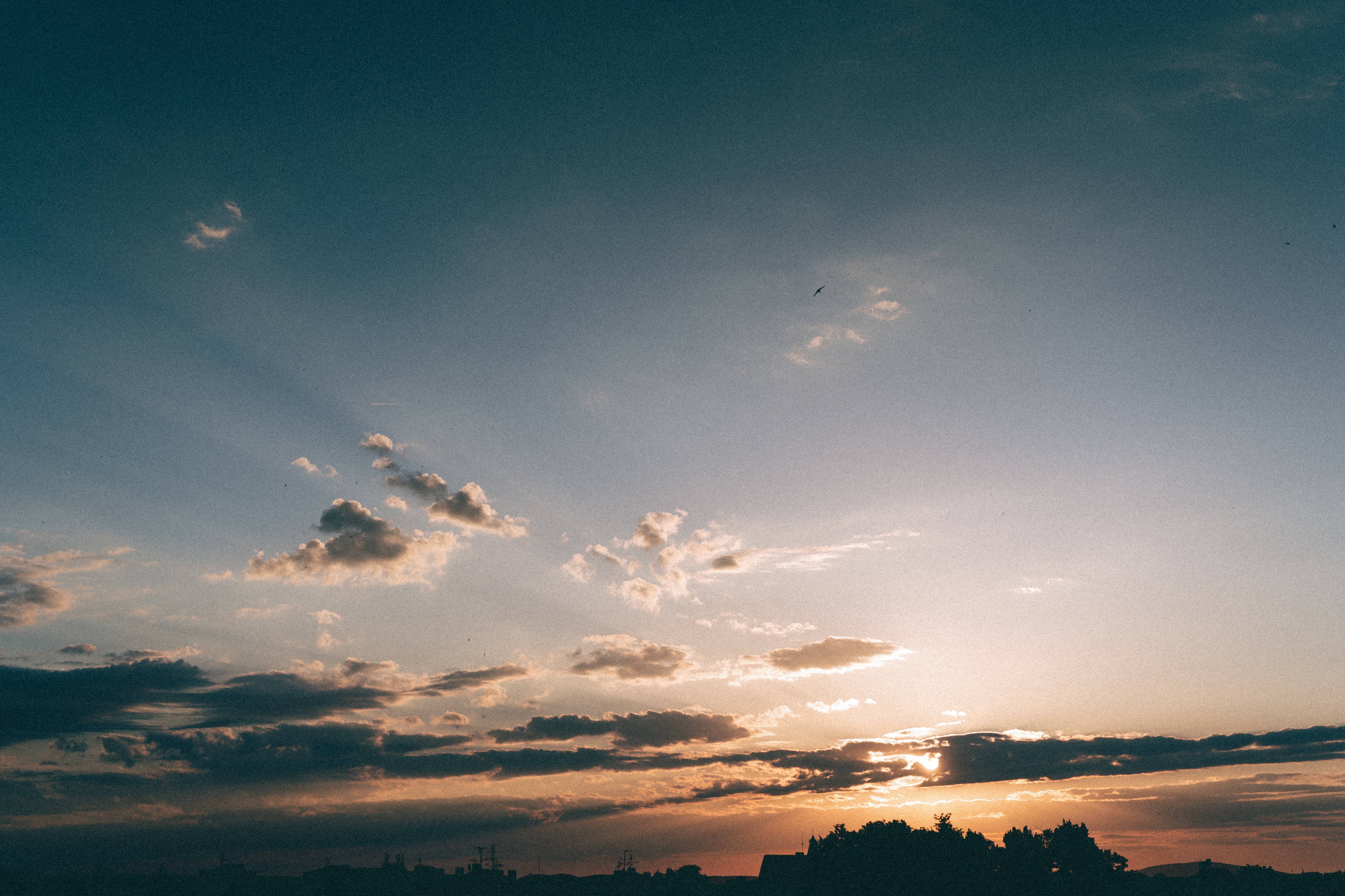 自然風景動圖 桌布 壁紙 — 夕陽的光線穿過散落的雲層，將金色的光芒投射在剪影般的樹木和遠處的建築物上。