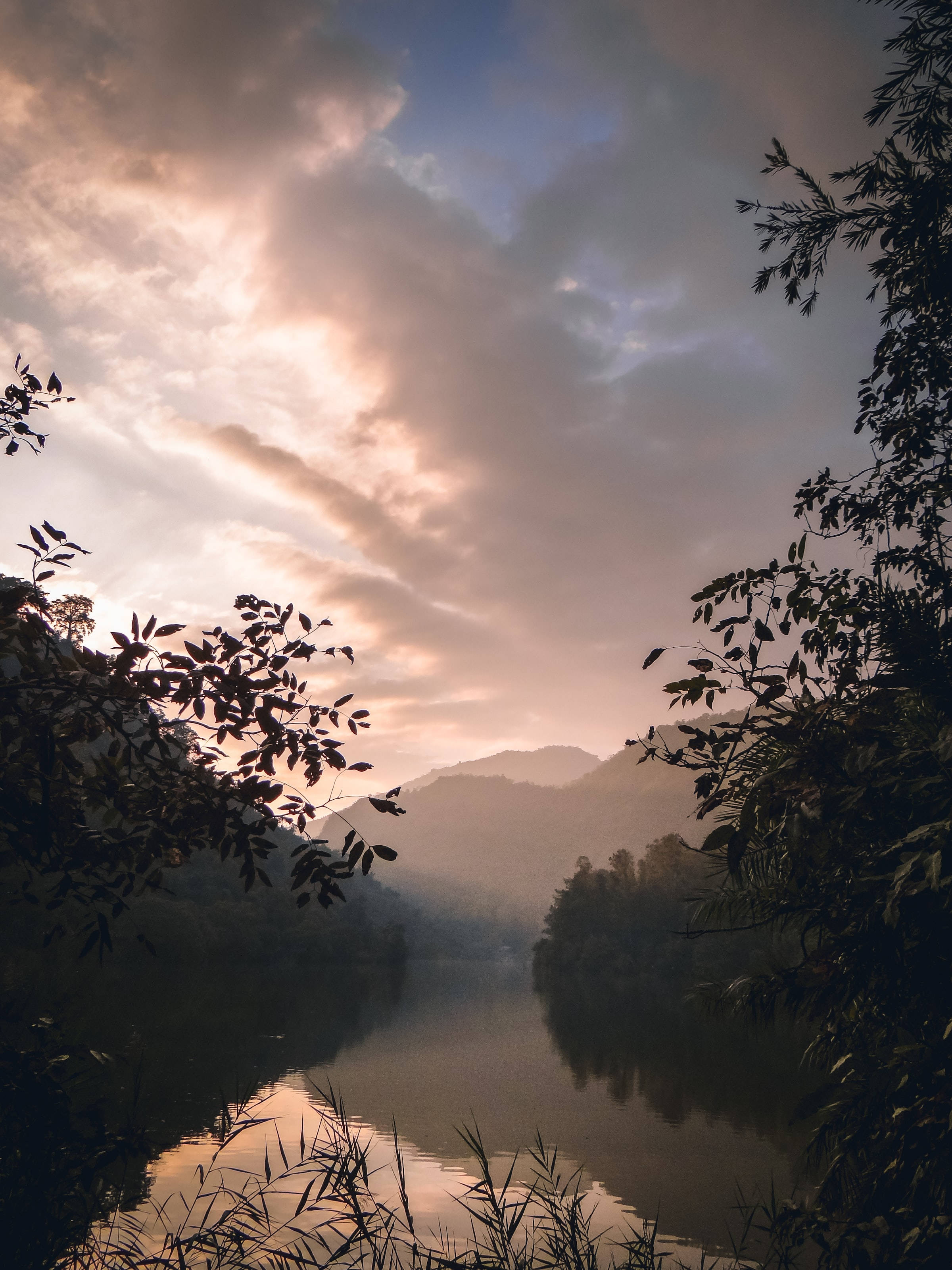 Lake 桌布 壁紙 — 夕陽的光線穿過雲層，灑在一條被樹枝剪影和遠山環繞的霧氣瀰漫的河流上。