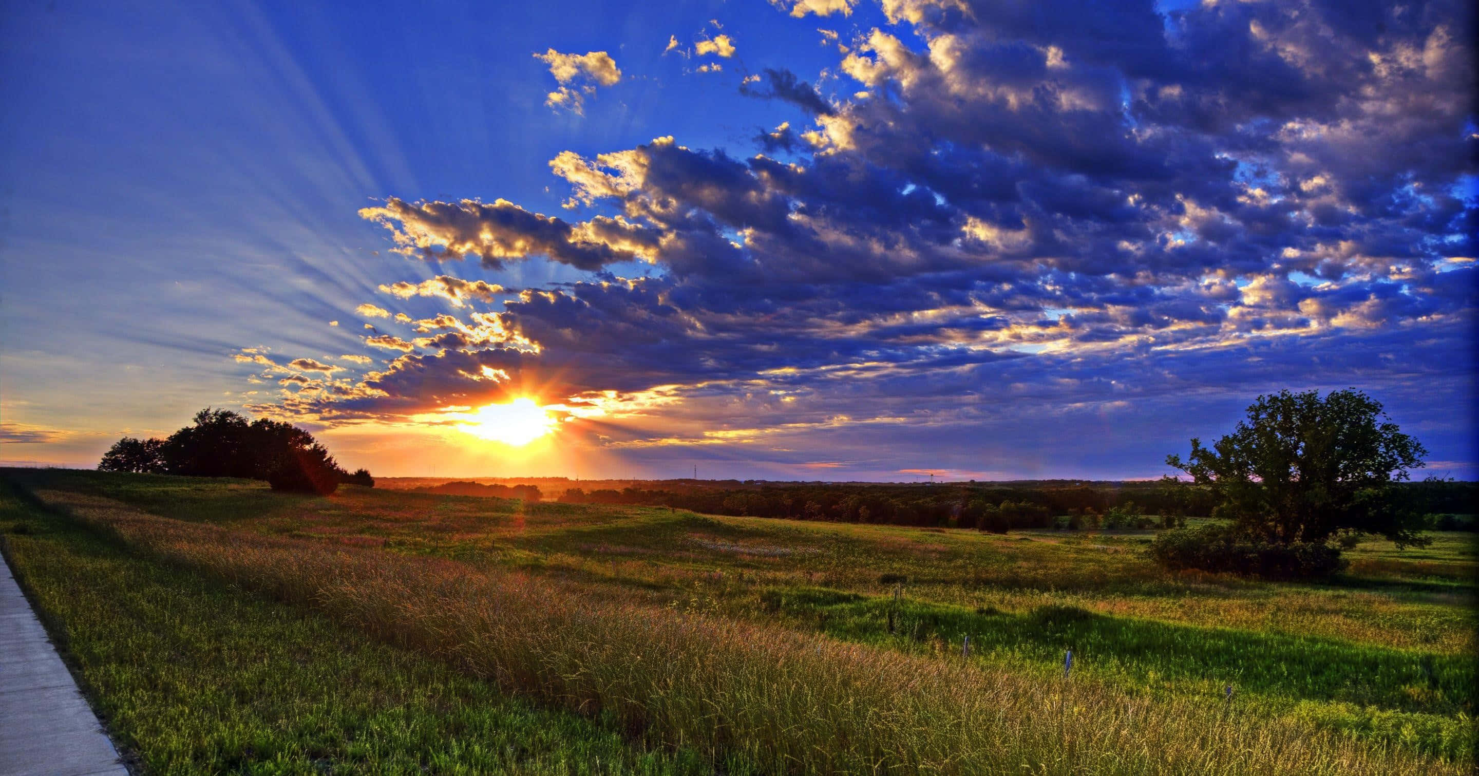 North Dakota 桌布 壁紙 — 夕陽穿透戲劇性的雲層，照耀著一片有散落樹木和蜿蜒小徑的草地。