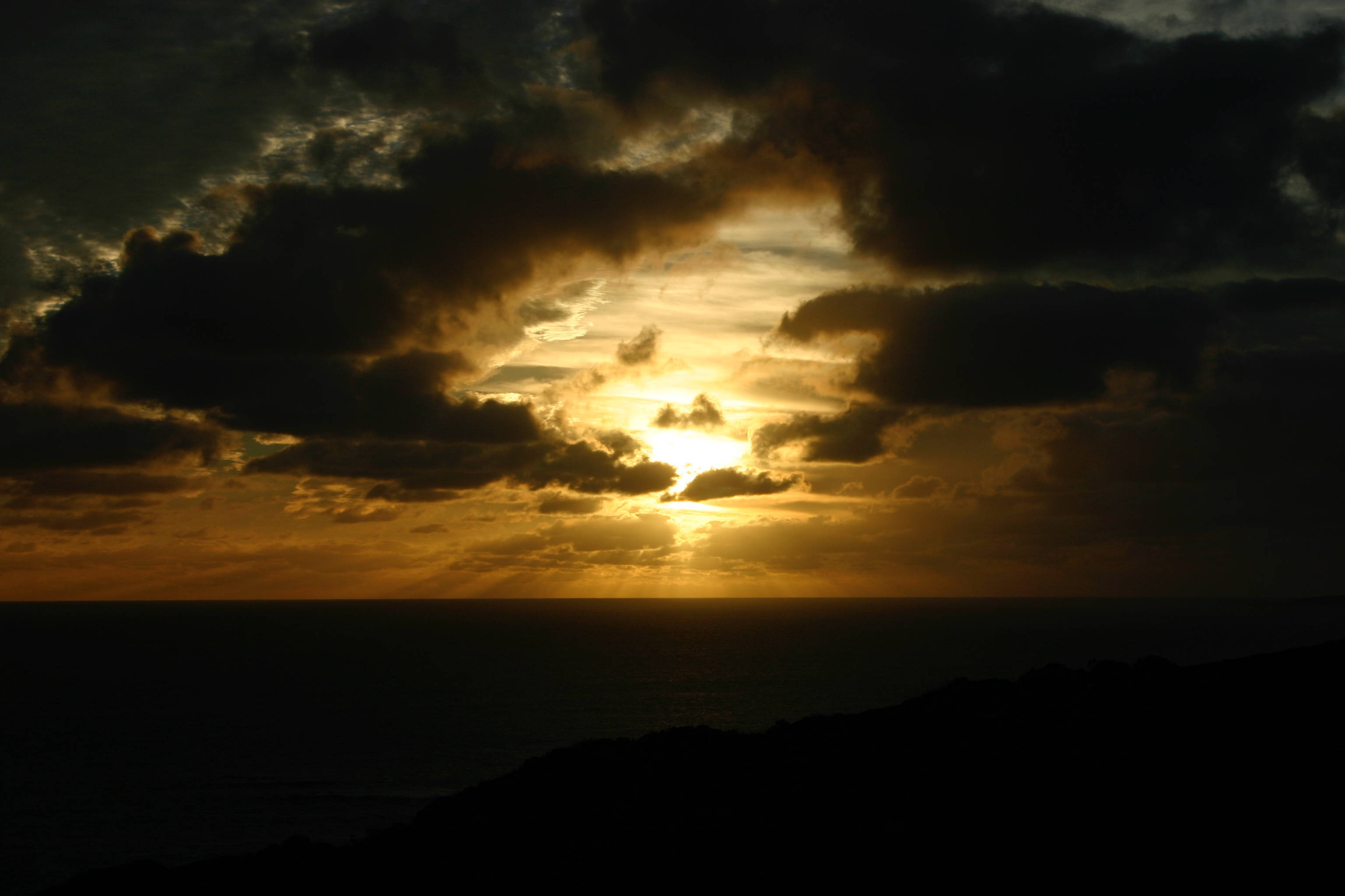 日落 桌布 壁紙 — 夕陽穿透暗雲，照耀在平靜的海洋上，下方是輪廓分明的山丘。