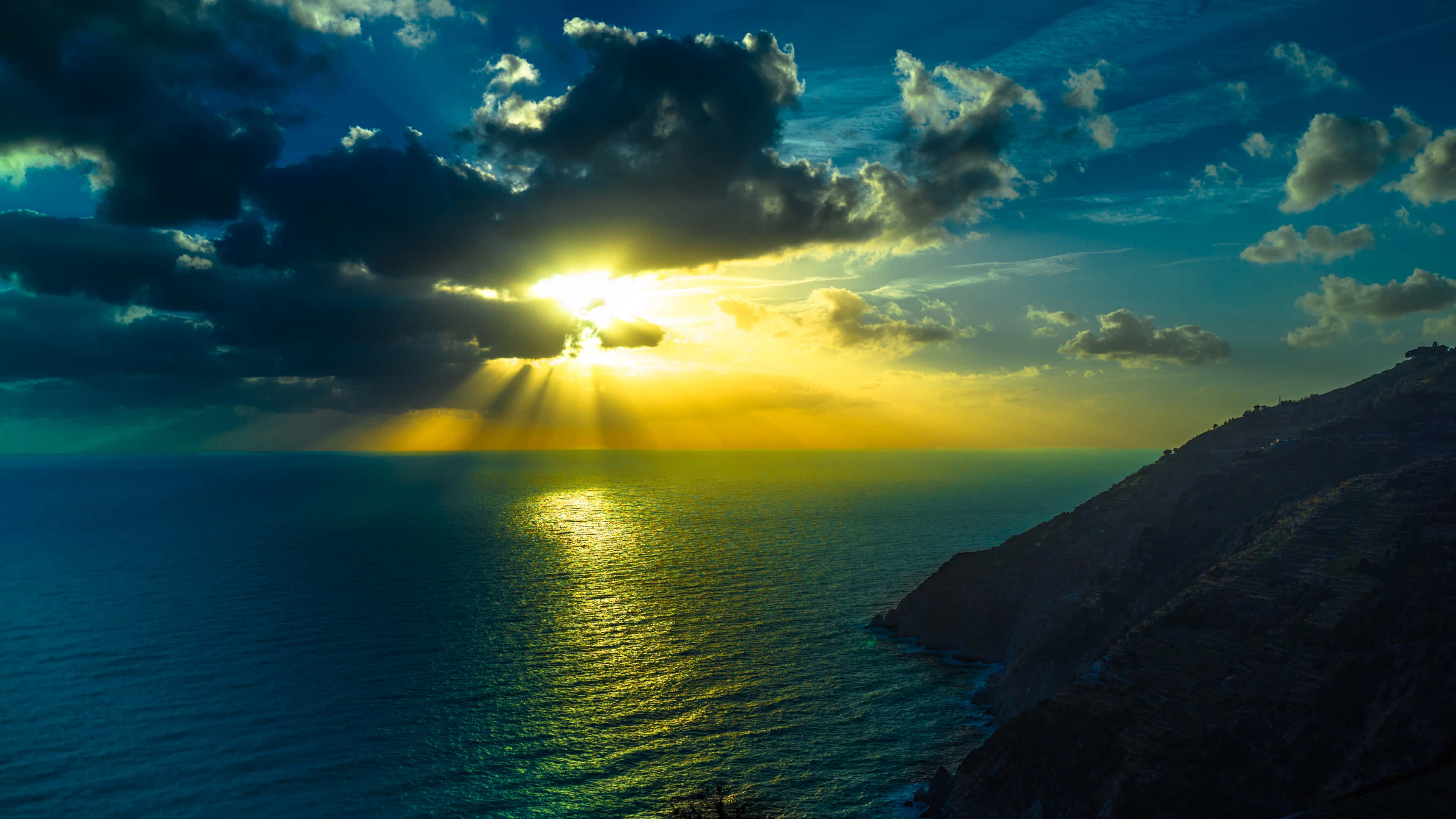 Ocean Sunset 桌布 壁紙 — 夕陽穿透深藍海洋旁崎嶇懸崖上方的烏雲。