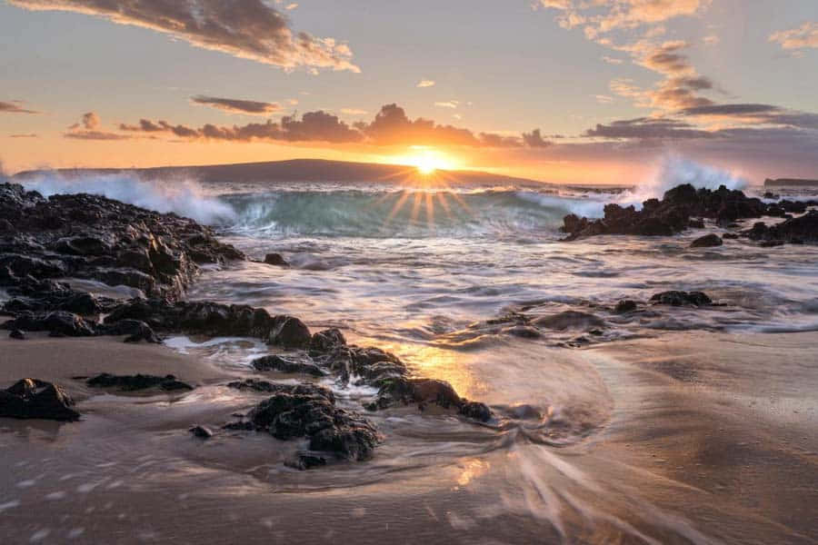 日落 桌布 壁紙 — 夕陽穿透雲層，海浪拍打沙灘上的深色火山岩，水花四濺。
