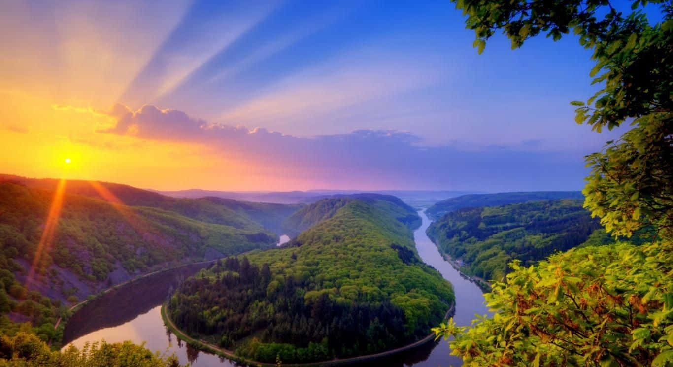 Beautiful Place 桌布 壁紙 — 夕陽穿透雲層，照耀在一條蜿蜒穿過翠綠山谷的河流上，前景的樹葉構成畫框。