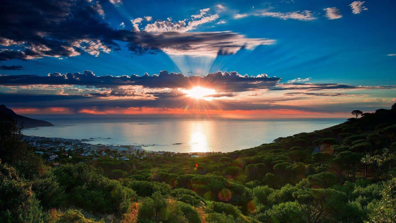 日落 桌布 壁紙 — 夕陽穿透雲層，照耀在坐落在翠綠山丘之間的海岸小鎮上。