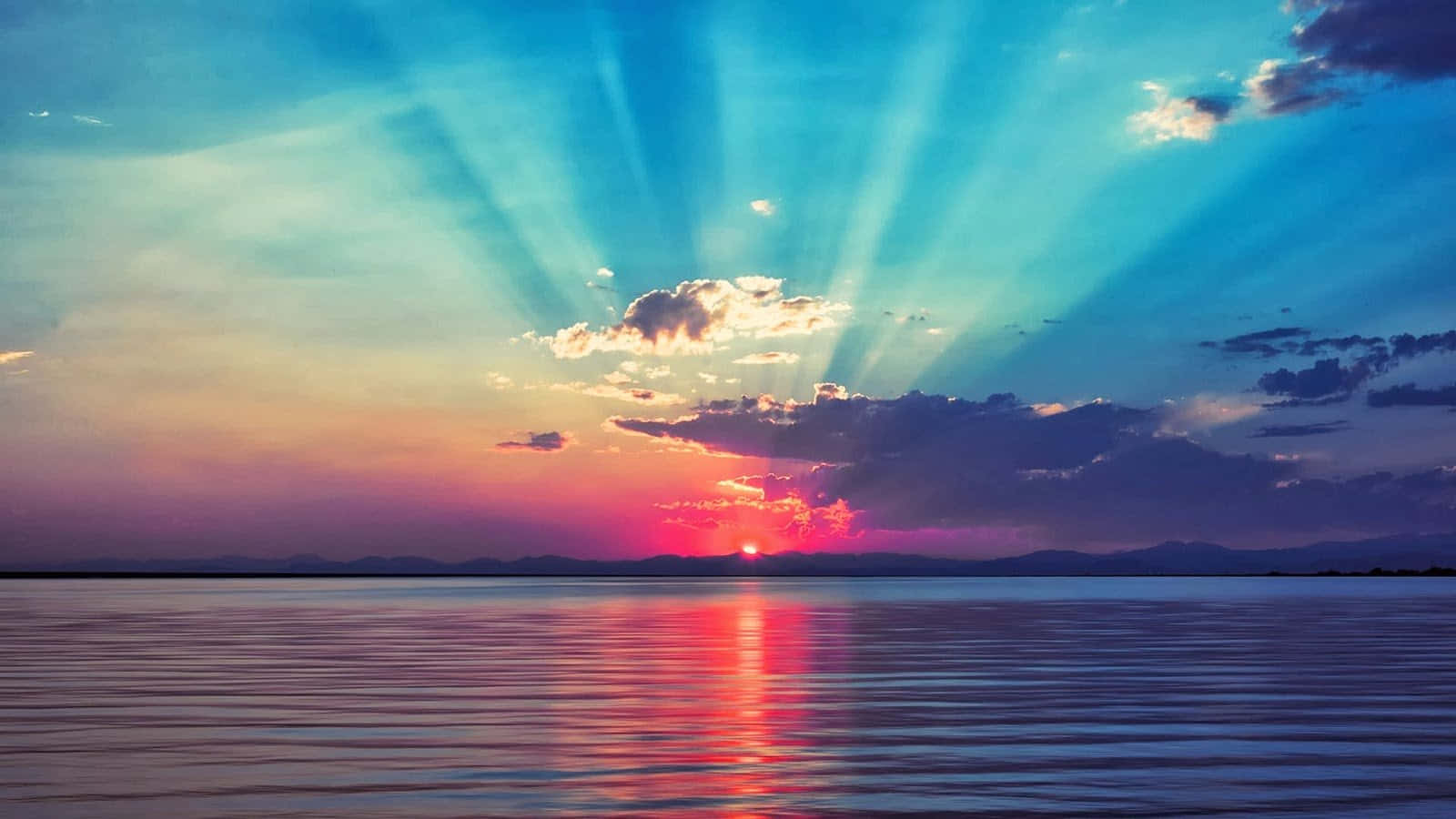 日出 桌布 壁紙 — 夕陽穿透雲層，照耀在平靜的水面上，倒映出火紅的粉藍色天空和遠山。