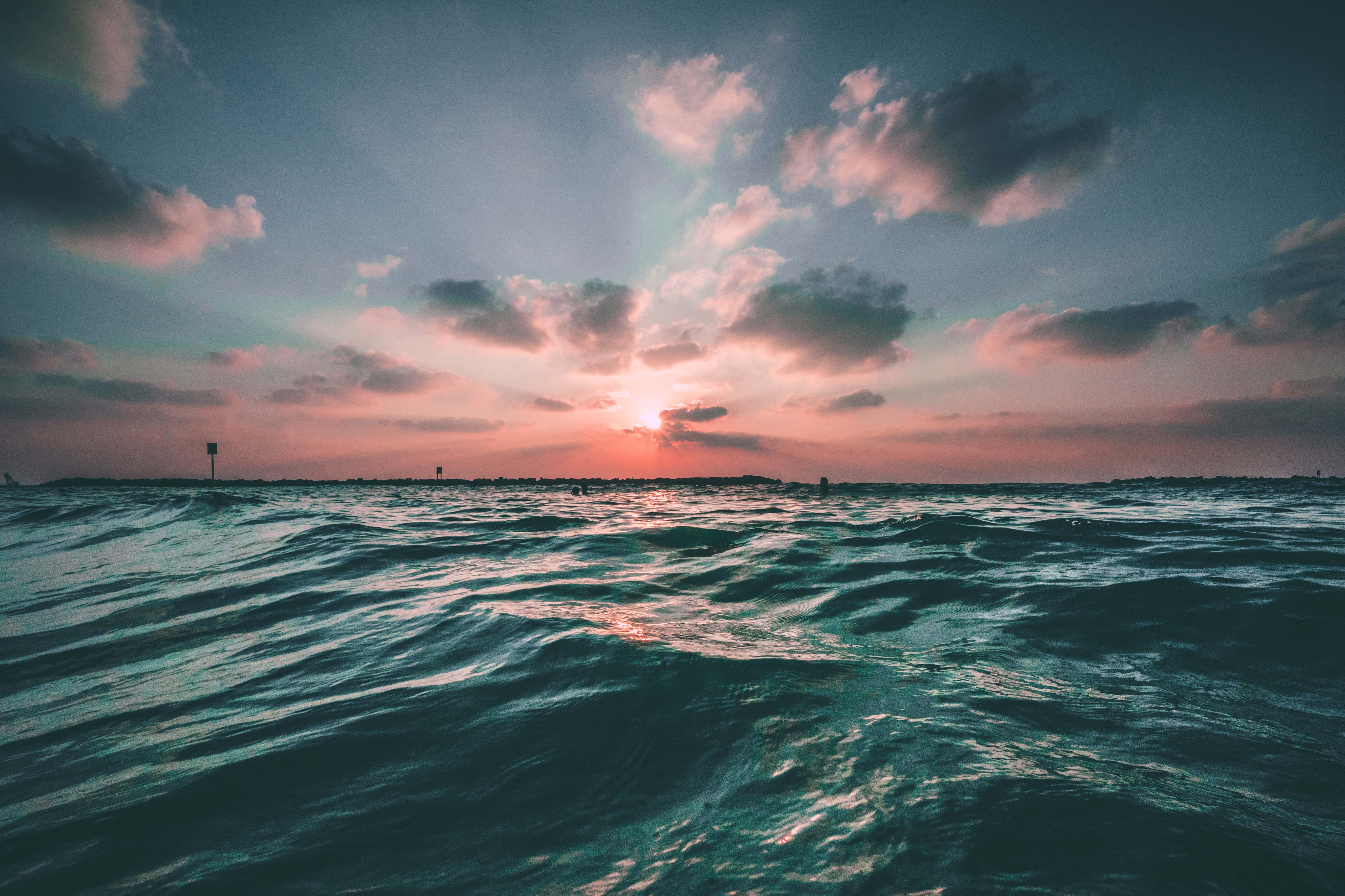 Cute Sea 桌布 壁紙 — 夕陽穿透雲層，照耀在波光粼粼的碧綠海洋上，地平線上有遠處的剪影。