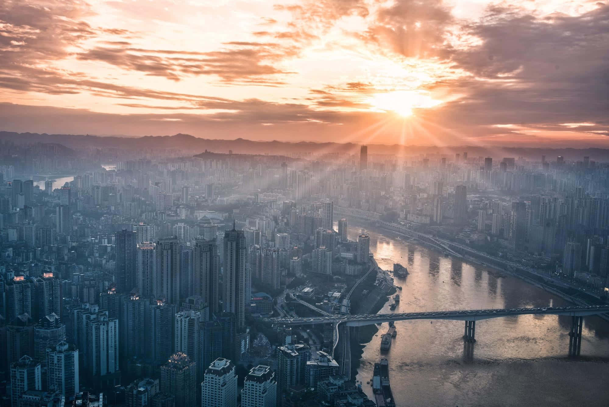 城市 桌布 壁紙 — 夕陽穿透雲層，照耀著蜿蜒河流和橋樑的廣闊城市景觀。