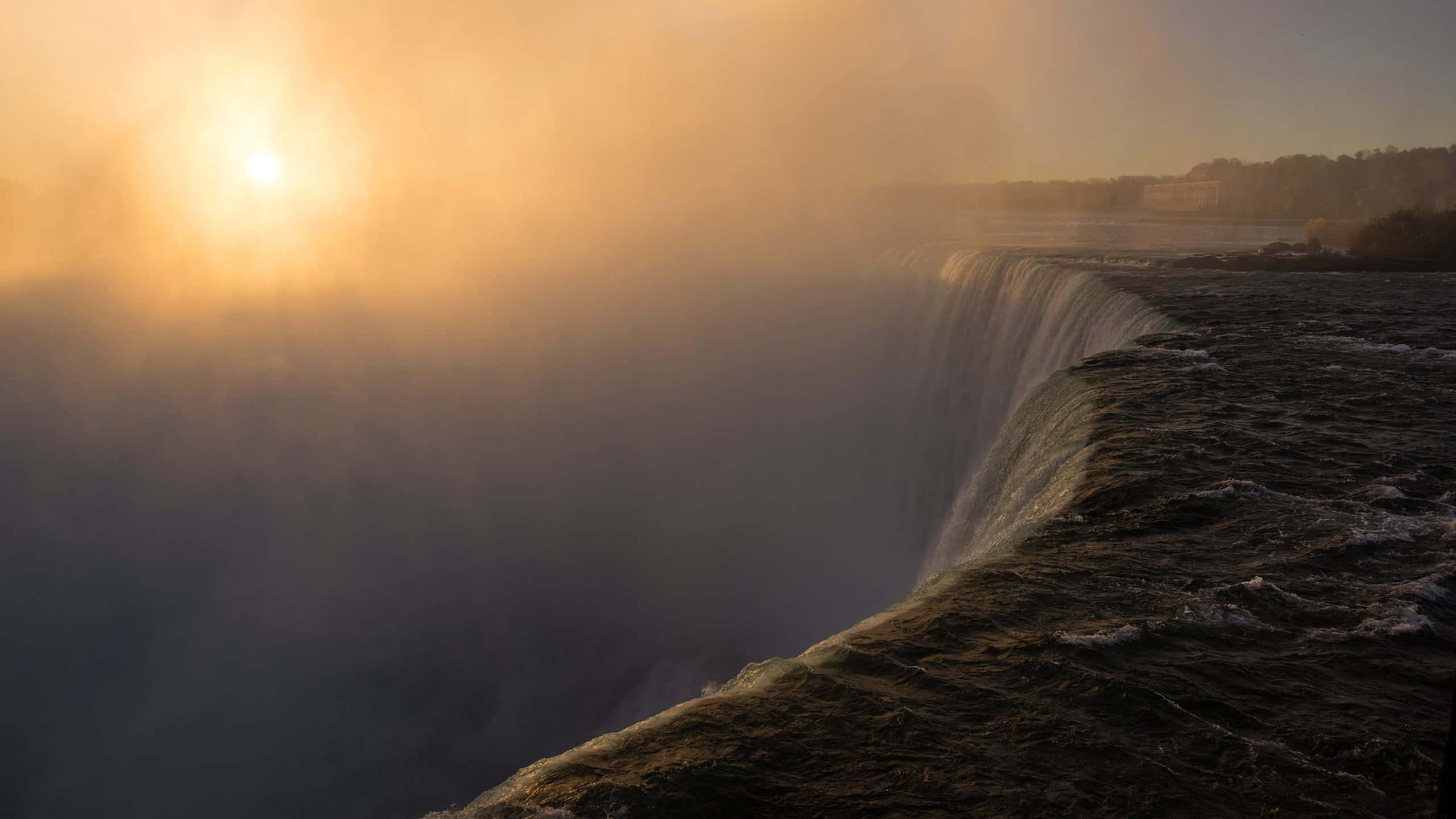 720p Niagara Falls 桌布 壁紙 — 夕陽穿過從巨大瀑布邊緣升起的薄霧，發出光芒，將金色的光線投射在奔流的水面上。