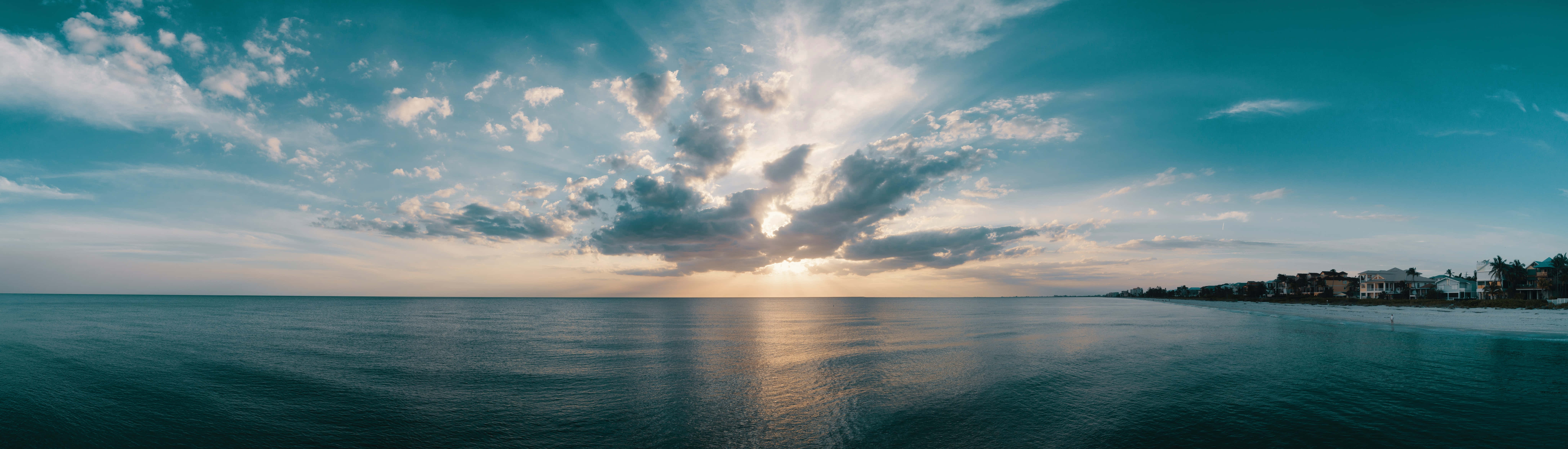 雙螢幕桌布 桌布 壁紙 — 夕陽穿過戲劇性的雲層，照耀在平靜的海洋上，遠處有房屋和棕櫚樹的海岸線。