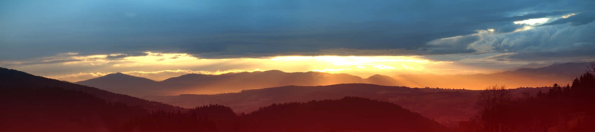 日落 桌布 壁紙 — 夕陽穿過暴風雲層，照耀在層層疊疊的山脊上，為森林覆蓋的山谷投下熾熱的橙色光芒。