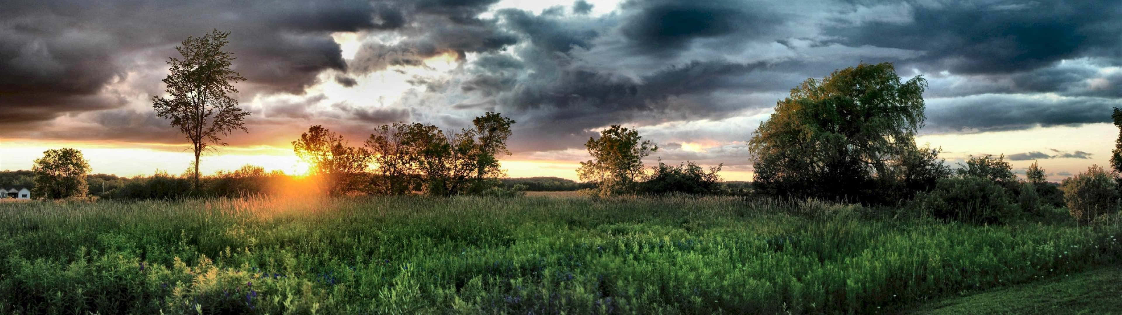 3840X1080 桌布 壁紙 — 夕陽穿過暴風雲層照耀，映照在廣闊的綠色田野上，點綴著樹木。