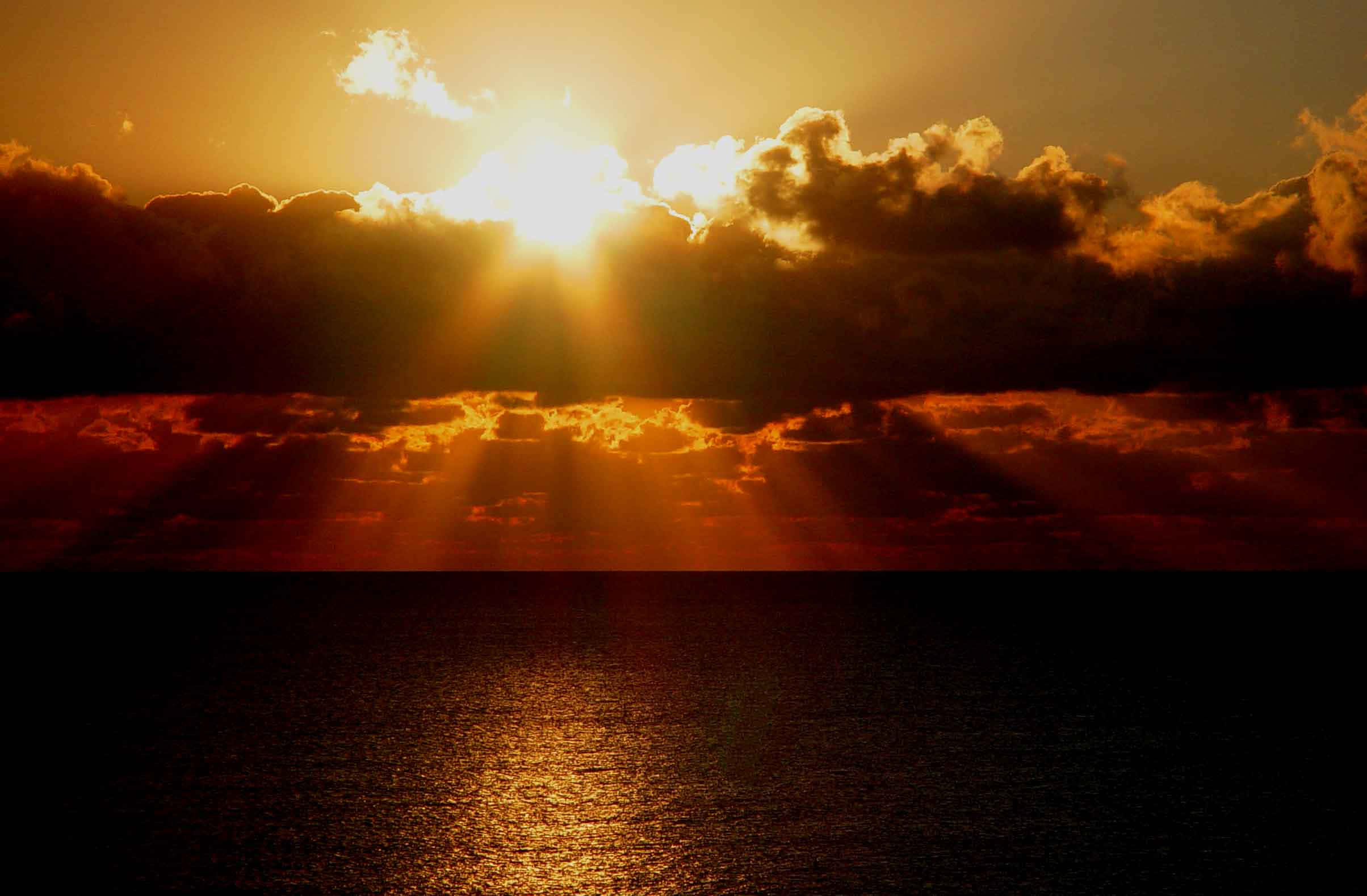 日落 桌布 壁紙 — 夕陽穿過烏雲，照射在平靜的海面上，反射出火橙色和深紅色的色調。