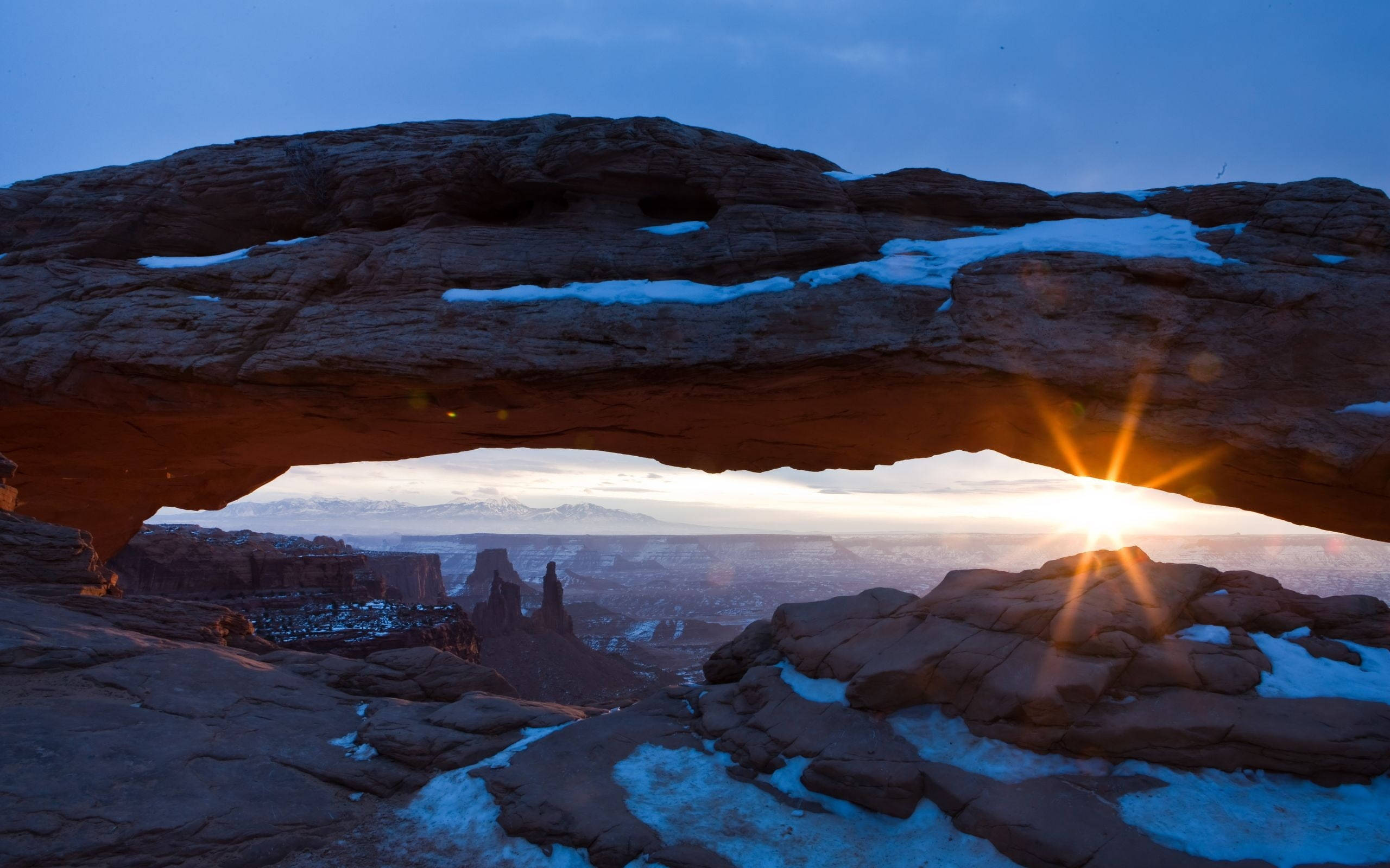 Canyonlands National Park 桌布 壁紙 — 夕陽穿過覆蓋著白雪的紅色岩石拱門，映襯著廣闊的峽谷景觀。