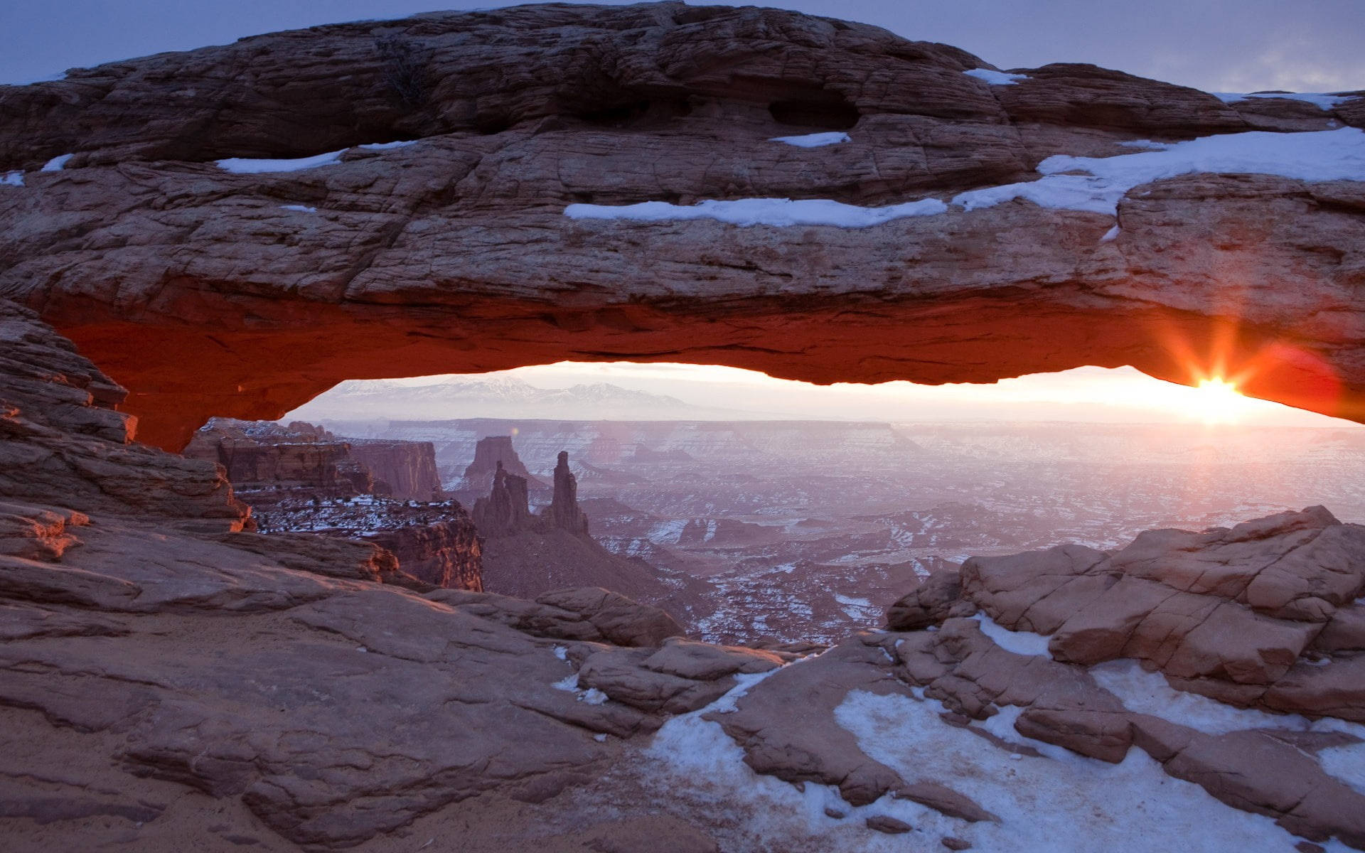 Canyonlands National Park 桌布 壁紙 — 夕陽穿過覆蓋著雪的紅色岩石拱門，映照出廣闊的峽谷景觀。