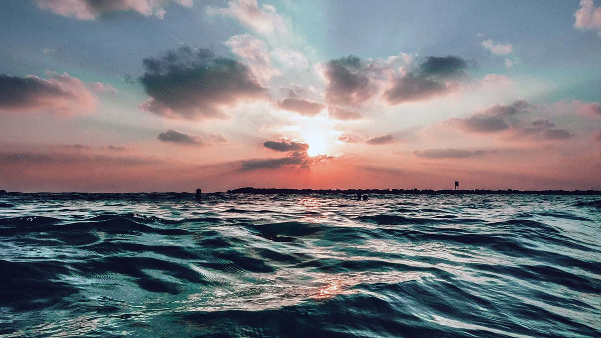 海洋 桌布 壁紙 — 夕陽穿過雲層，照射在紋理豐富的海洋上，遠方有陸地輪廓。