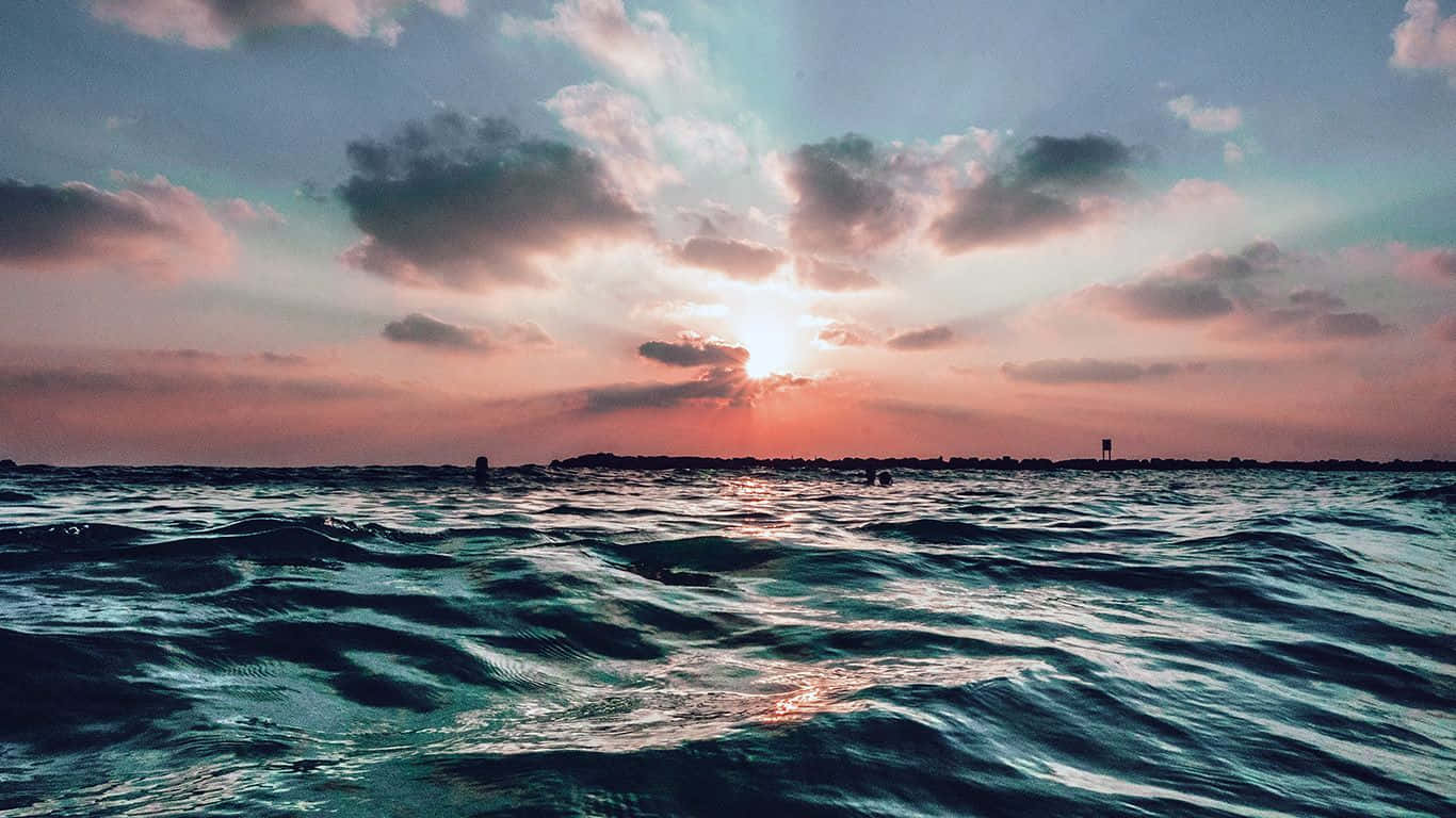 海灘 桌布 壁紙 — 夕陽穿過雲層，照耀在紋理豐富的海洋上，遠處有陸地輪廓。