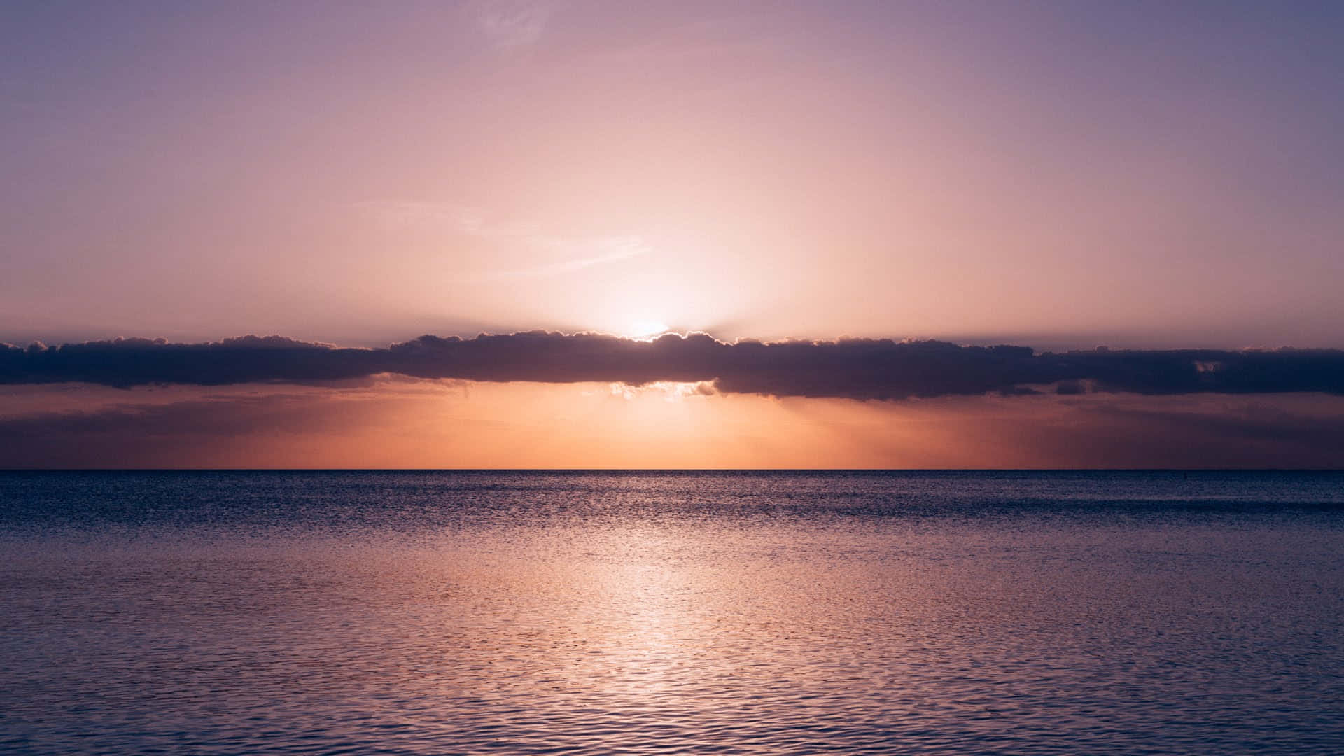 平靜 桌布 壁紙 — 夕陽穿過雲層發光，在黃昏時分將金色光芒投射在平靜的海面上。