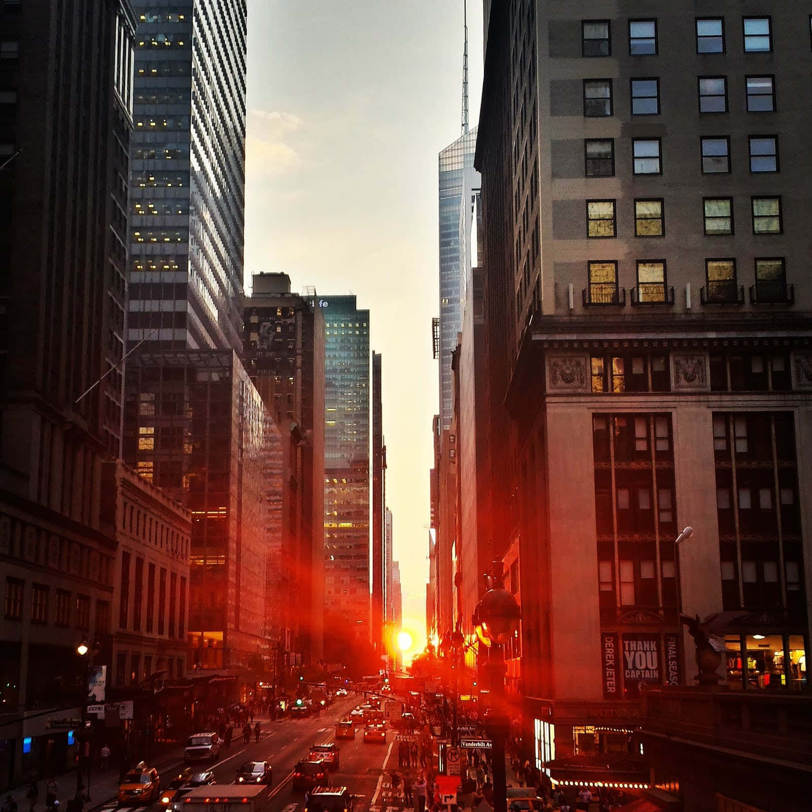 Cute Sunset 桌布 壁紙 — 夕陽穿過高聳的摩天大樓閃耀，將溫暖的光線投射在充滿汽車和行人的繁忙城市街道上。