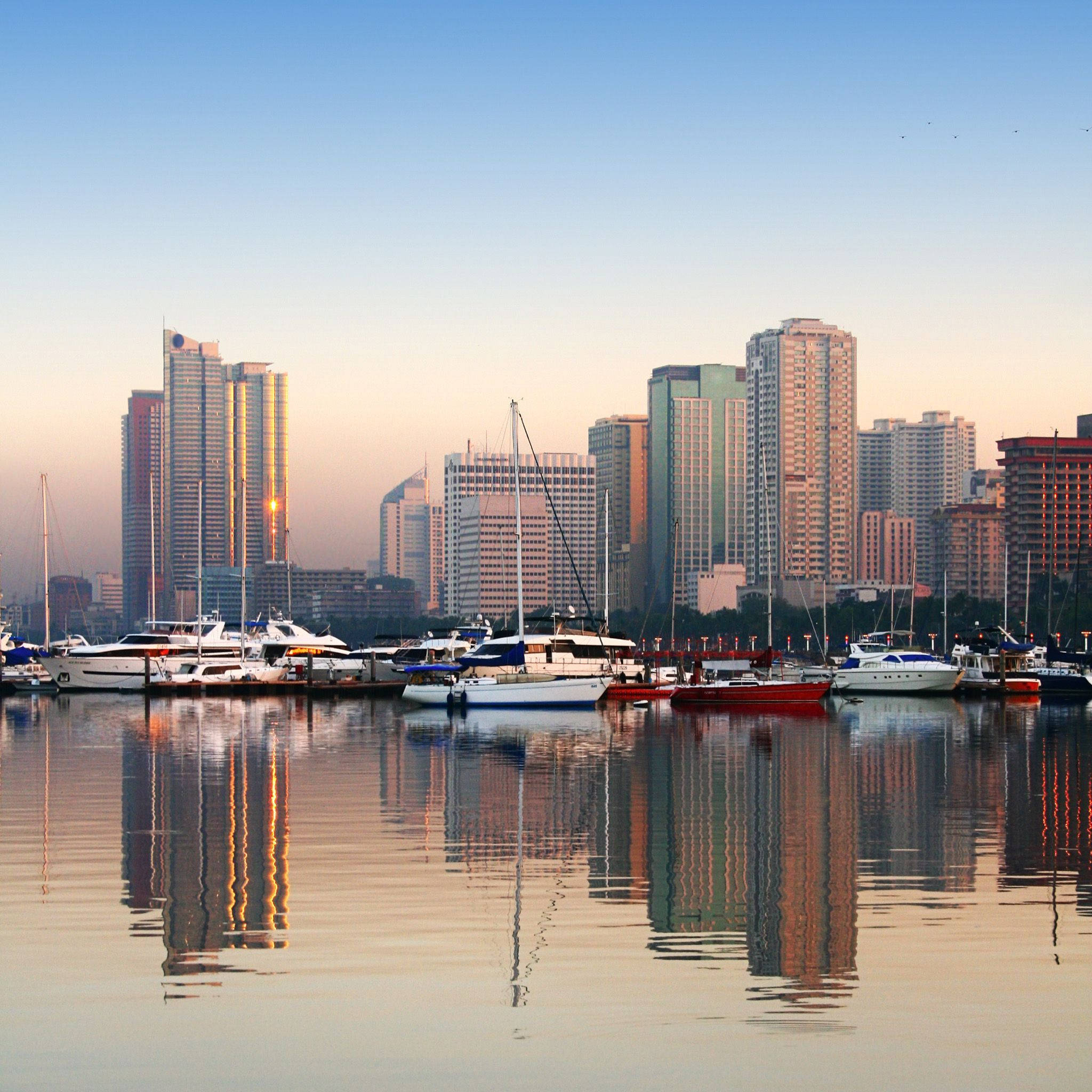 Manila 桌布 壁紙 — 夕陽色彩描繪出一個寧靜的港口，豪華遊艇和帆船在玻璃般的水面上倒映，背景是現代城市天際線。